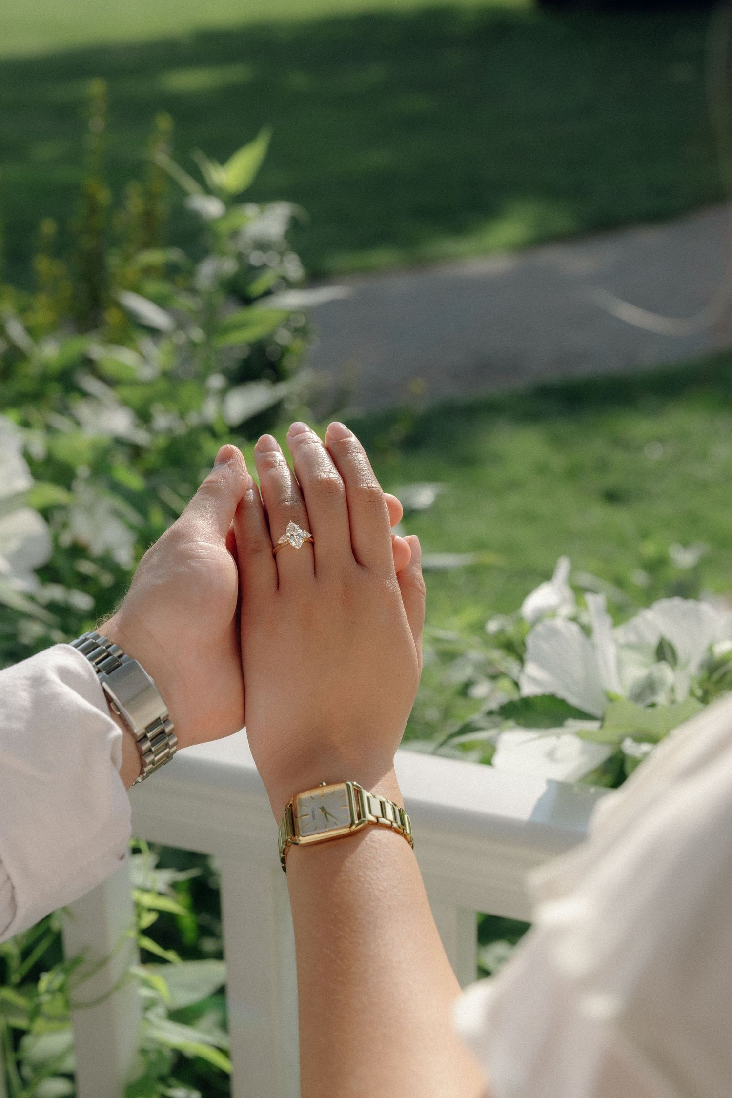 Two hands touching, one wearing a diamond engagement ring and gold watch, the other a silver watch, with green garden background.