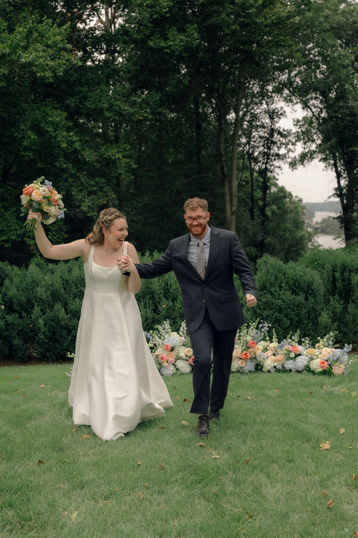 Joyful newlyweds walk across green lawn, bride in white gown holding bouquet, groom in dark suit, floral backdrop.
