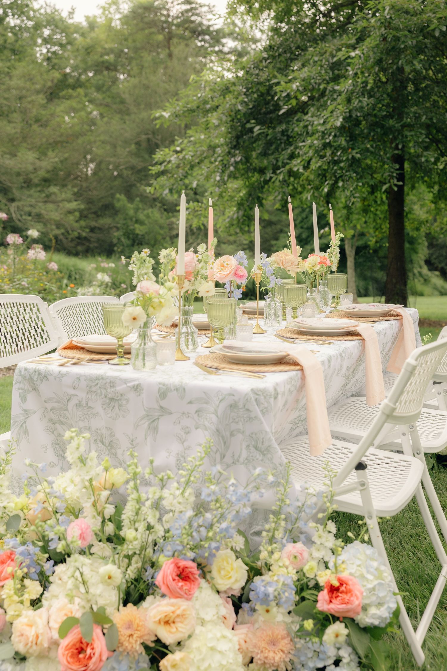 Elegant outdoor wedding table with floral tablecloth, candles, pastel blooms, and white chairs set in a lush garden.