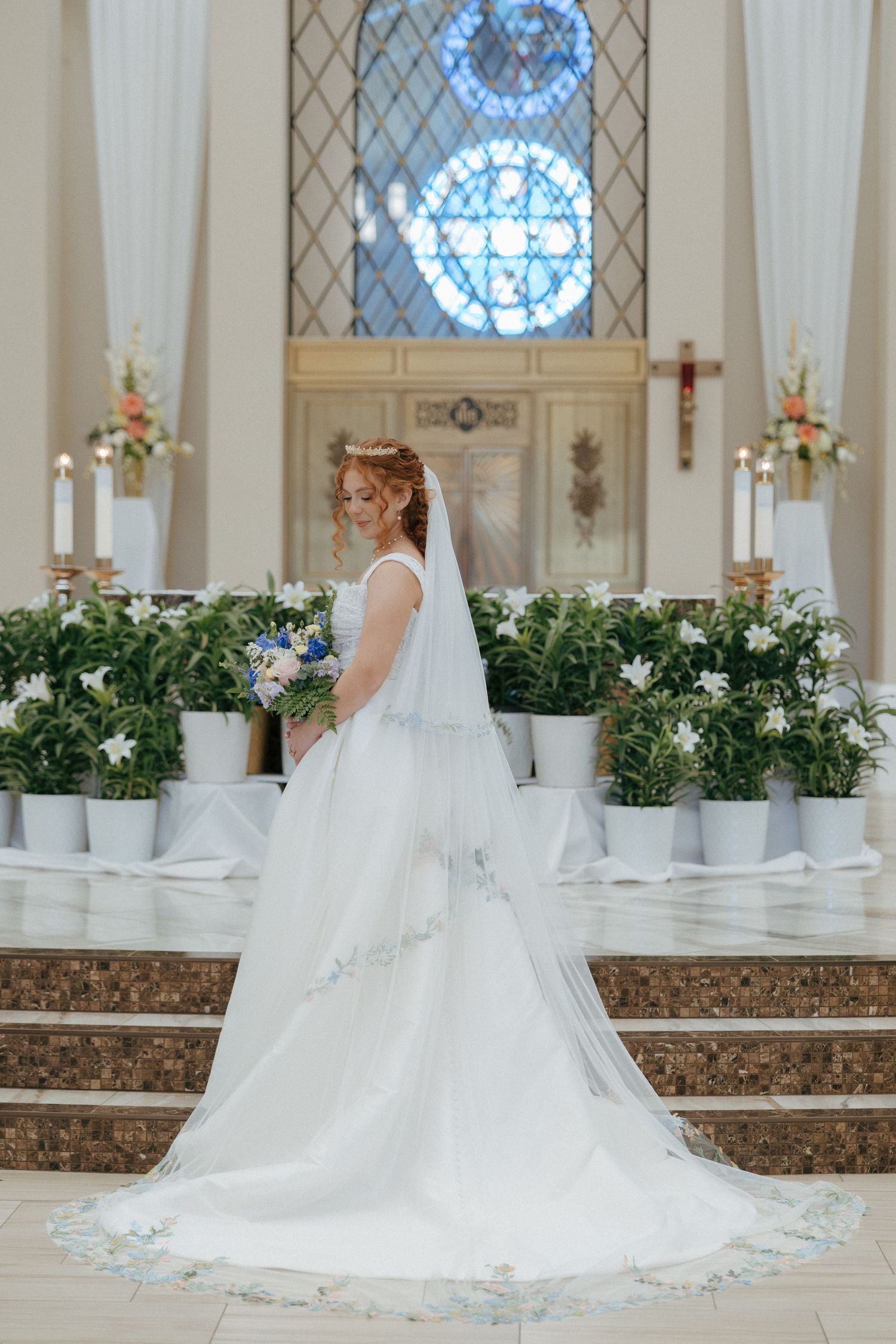 Bride in white gown and veil holds bouquet at church altar with stained glass window and floral arrangements.