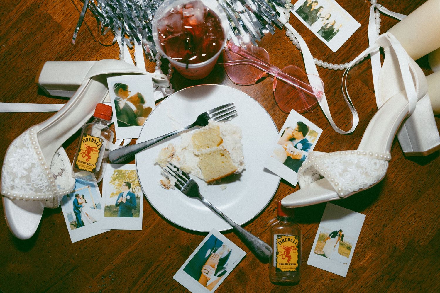 Flat lay of wedding shoes, Polaroid photos, cake slices, forks, and mini whiskey bottles on a wooden floor.