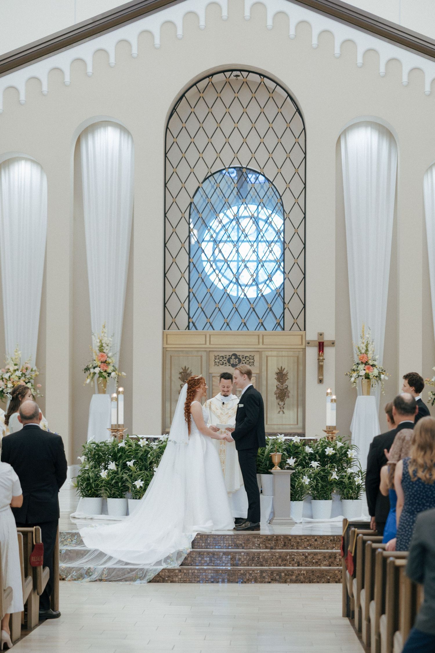 Bride and groom exchange vows at a elegant church altar with stained glass window, floral arrangements, and guests seated in pews.