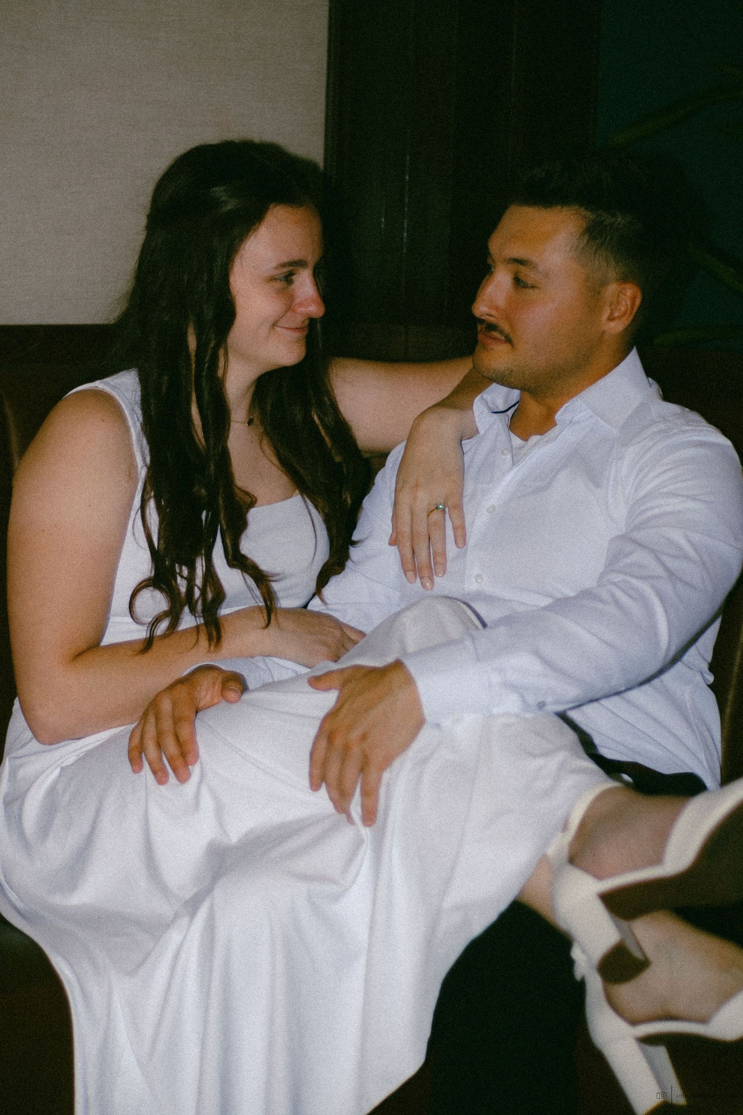 A couple in white attire sits together, sharing an intimate moment, with the woman smiling at the man.