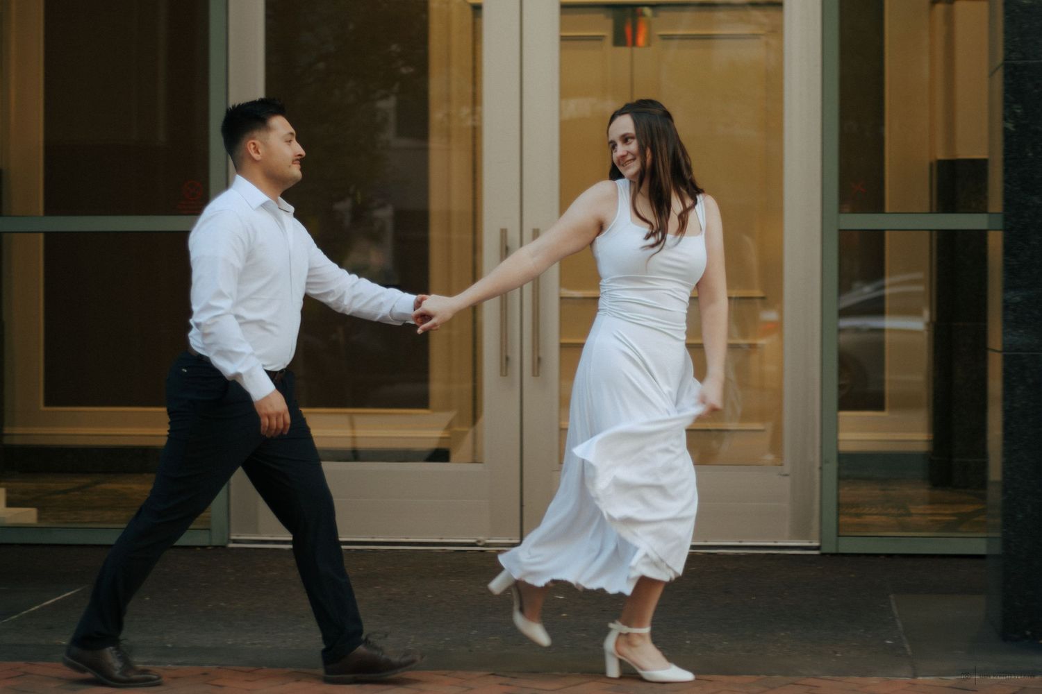 Joyful couple holding hands and dancing outside a building, woman in flowing white dress, man in white shirt.