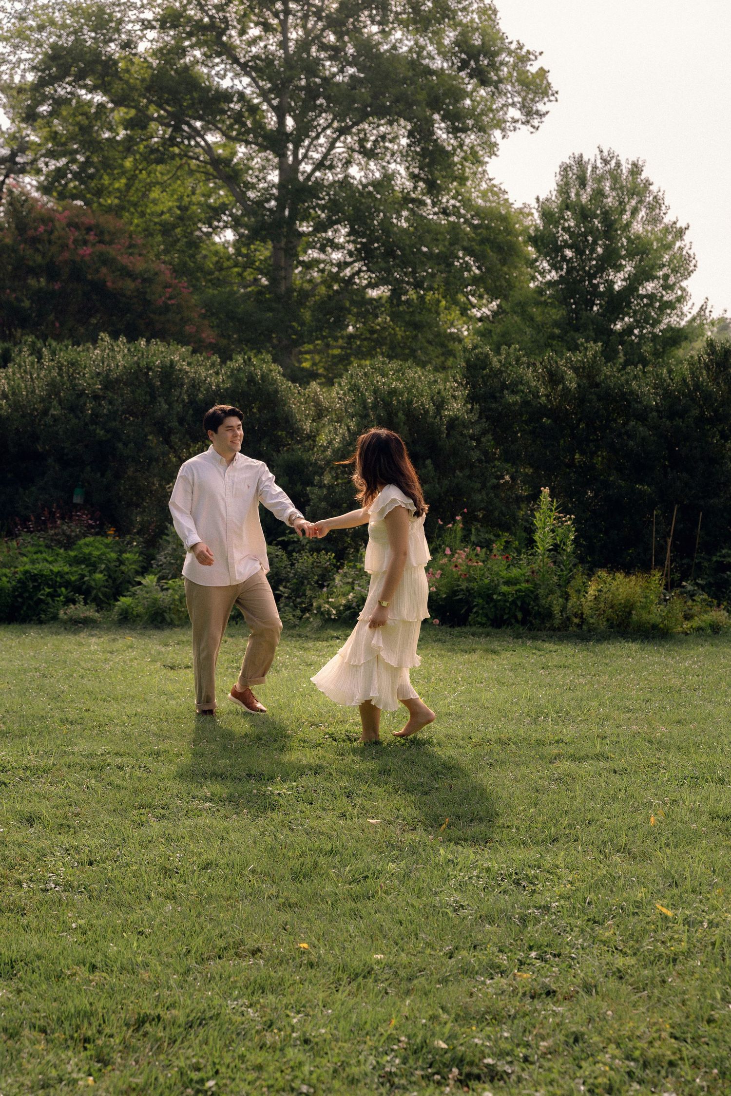 A couple holding hands and dancing joyfully on a green lawn surrounded by lush trees in a garden.