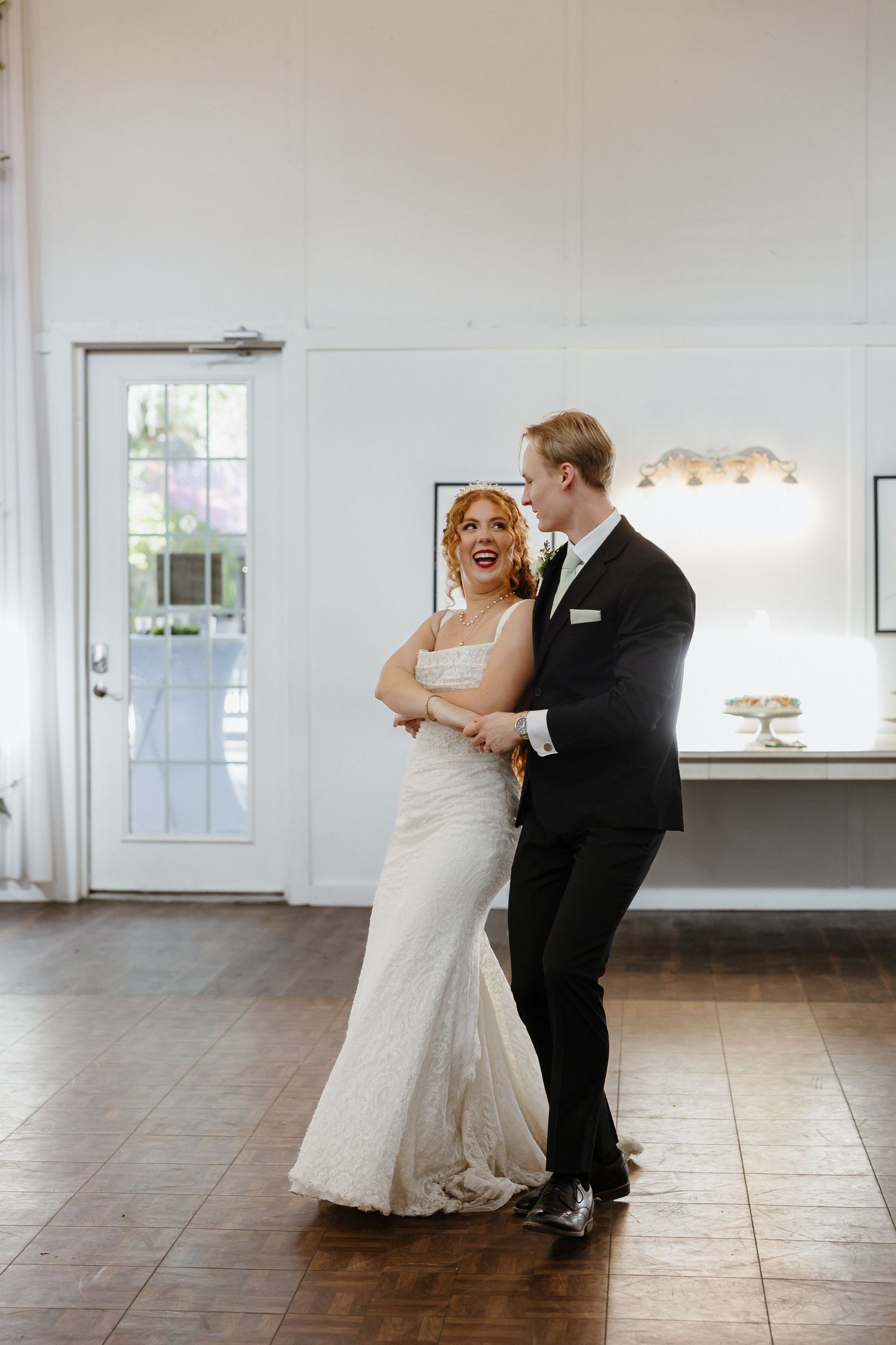 Bride and groom share their first dance in a bright white reception hall, bride in fitted lace gown, groom in classic black tuxedo.