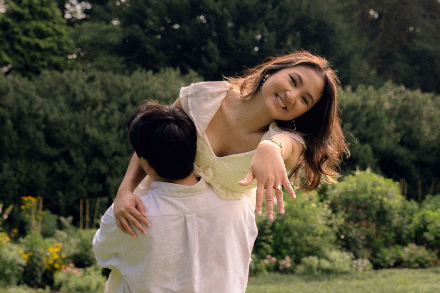 A joyful woman with long hair smiles while being lifted by a person in a lush green garden.