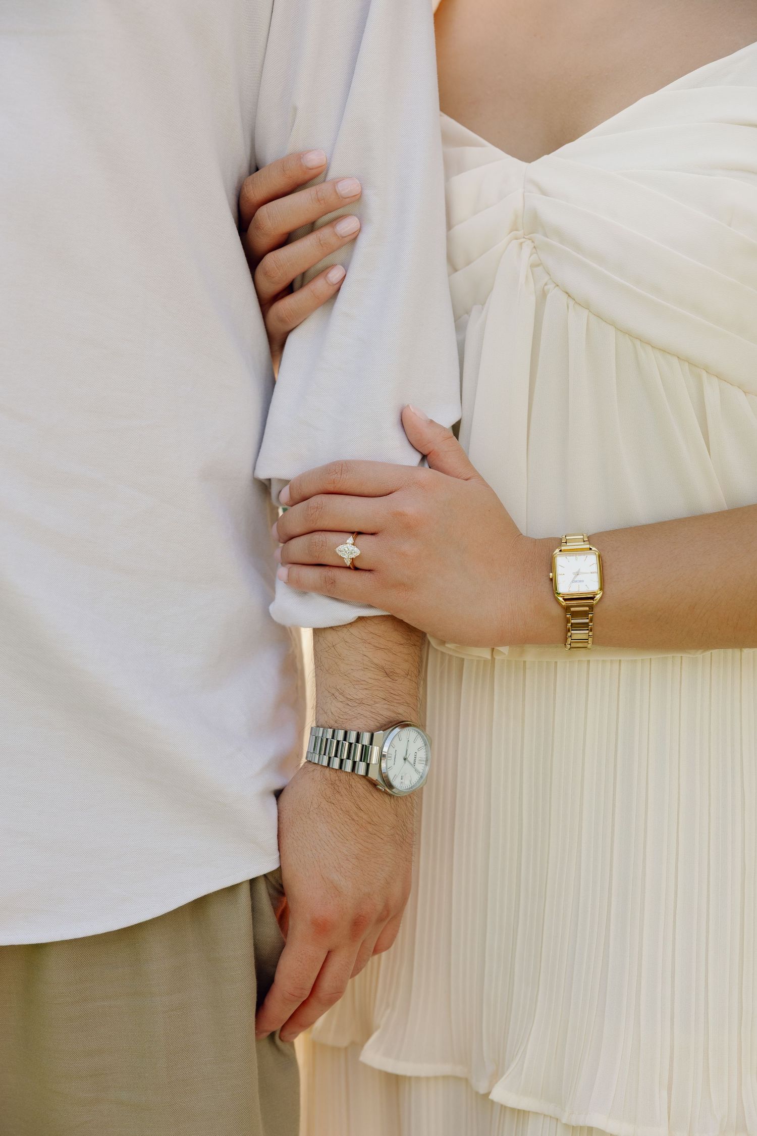 Couple in white outfits showing engagement ring and watches, woman in dress holding man's arm close.