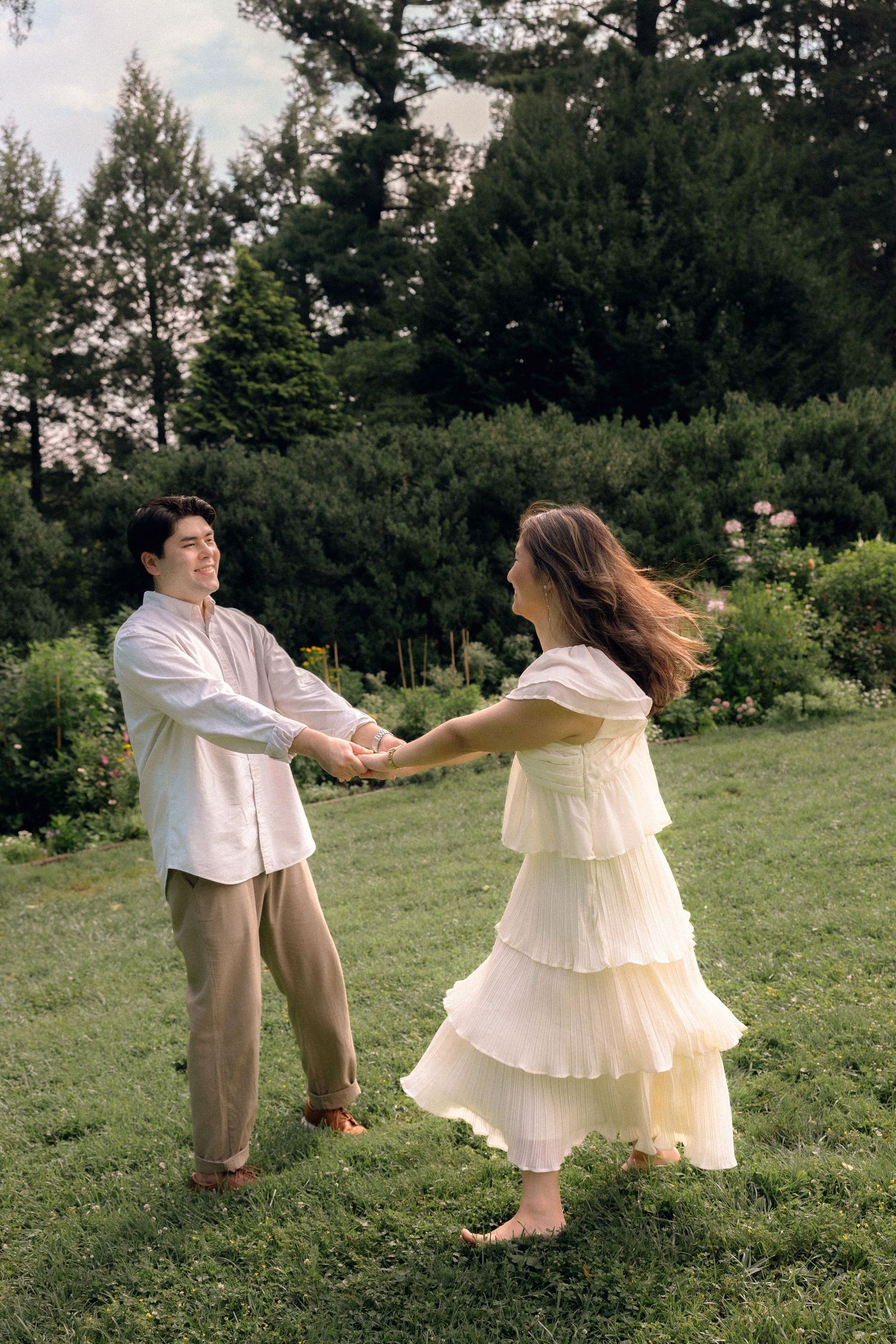 Couple holding hands and spinning on green grass in a garden, woman in flowing white tiered dress, man in white shirt and tan pants.