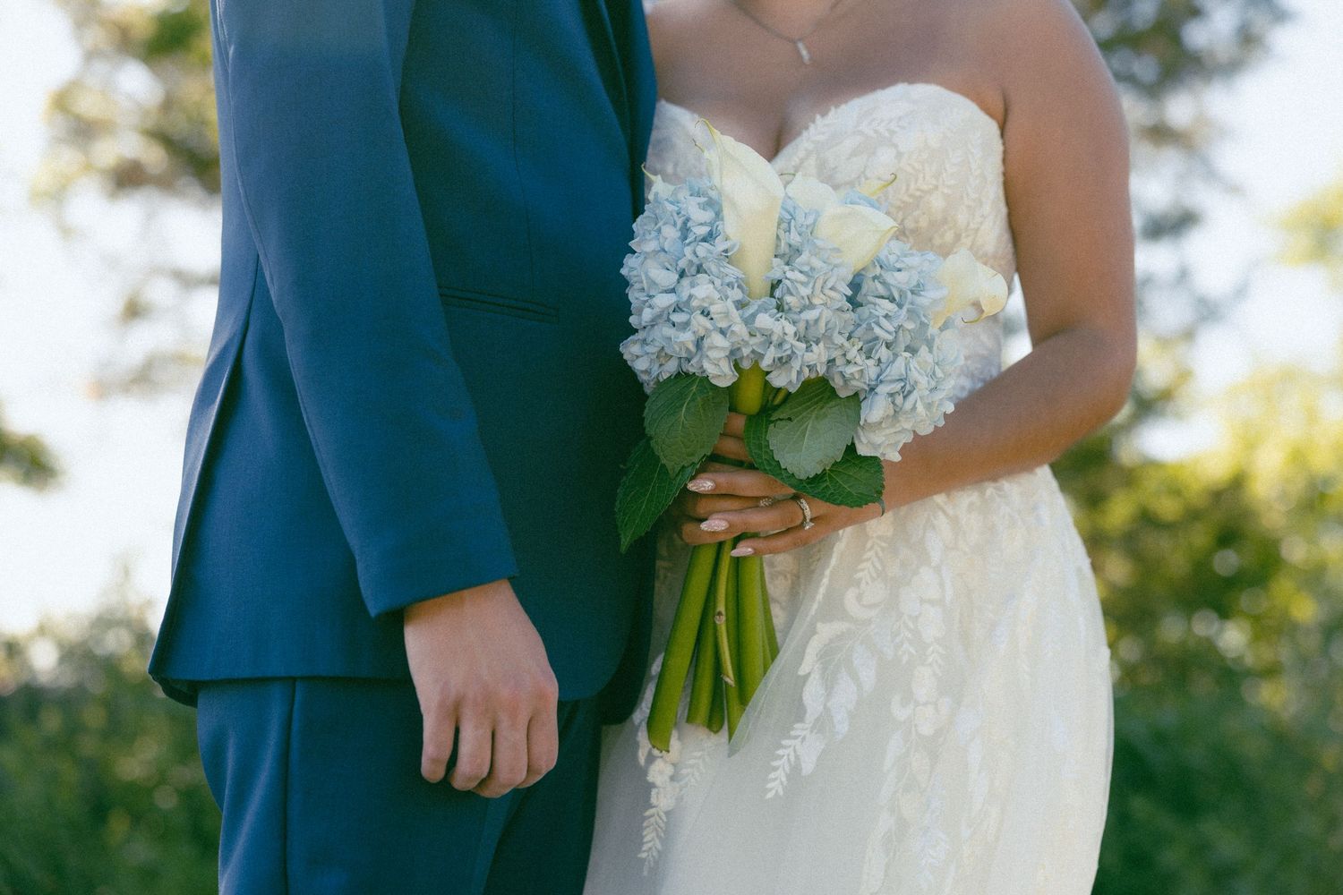 Bride in lace gown and groom in navy suit holding a blue hydrangea bridal bouquet outdoors.