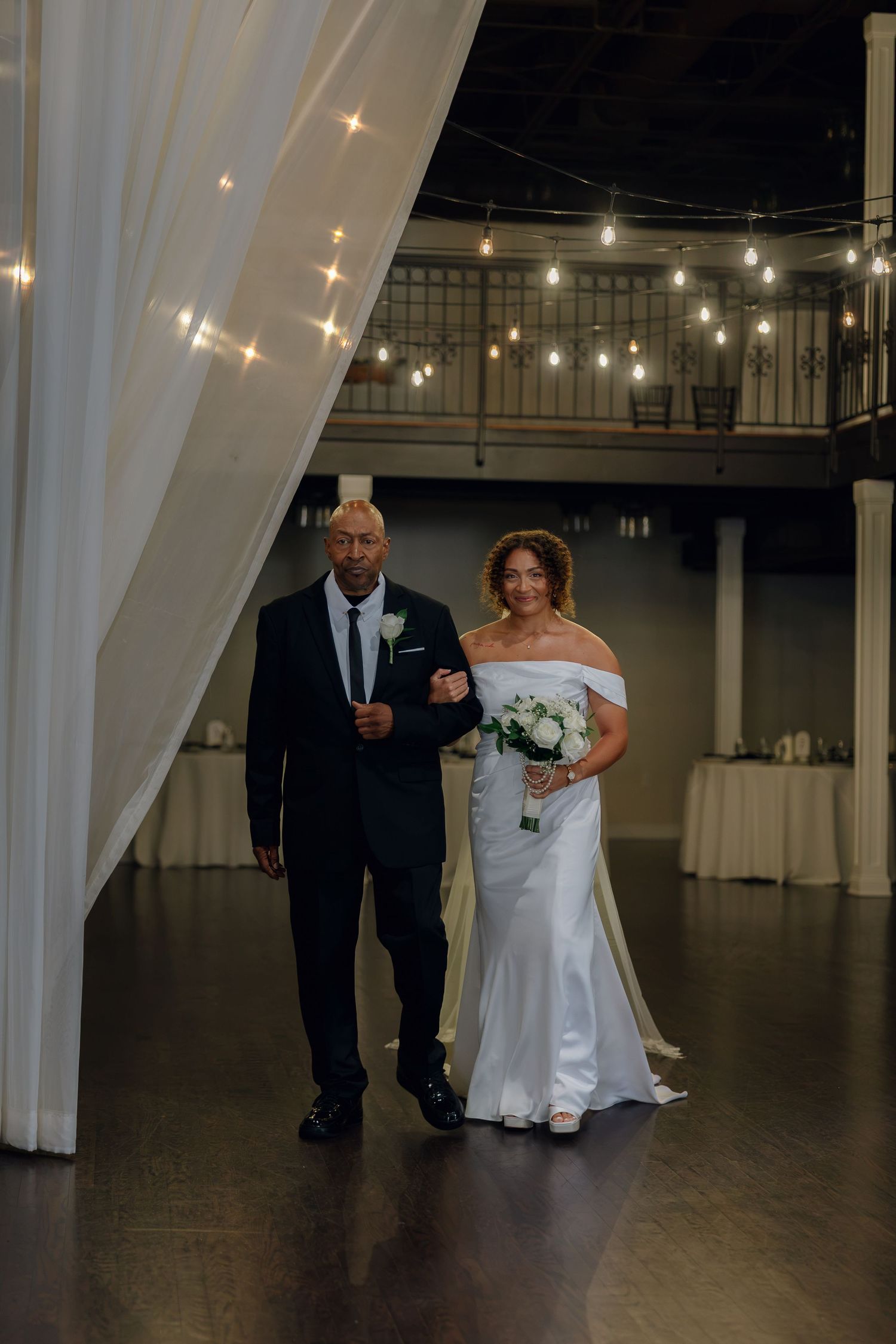 Bride in off-shoulder white gown holding bouquet walks with escort through draped entrance with string lights.