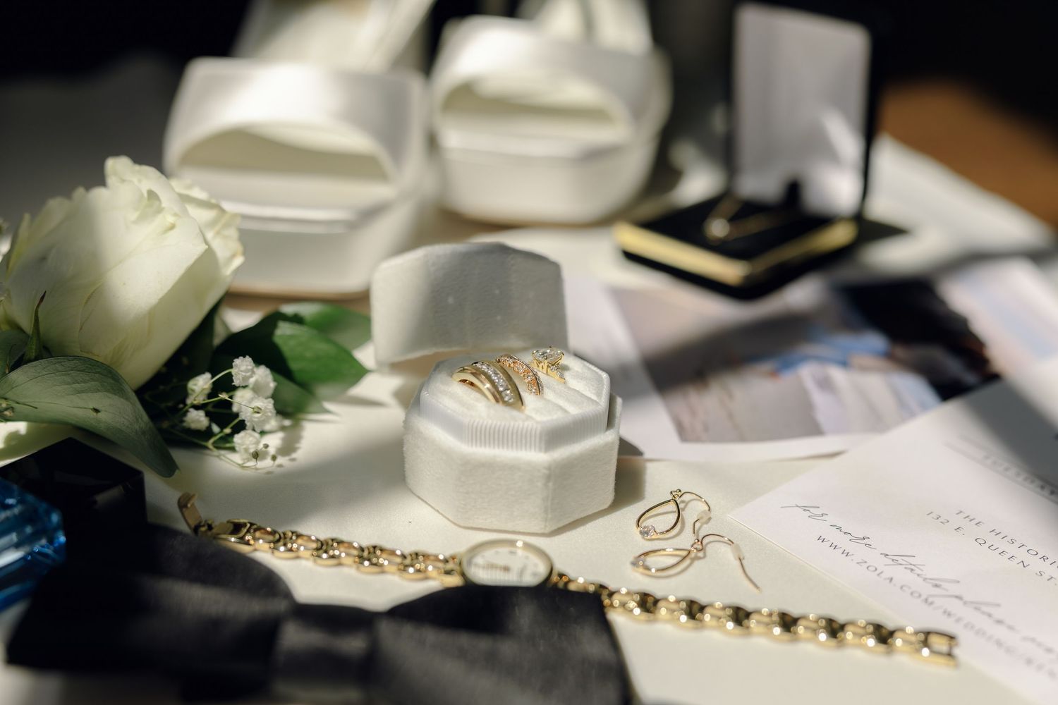Wedding rings in white velvet box surrounded by bridal accessories, gold bracelet, white roses, and elegant shoes.