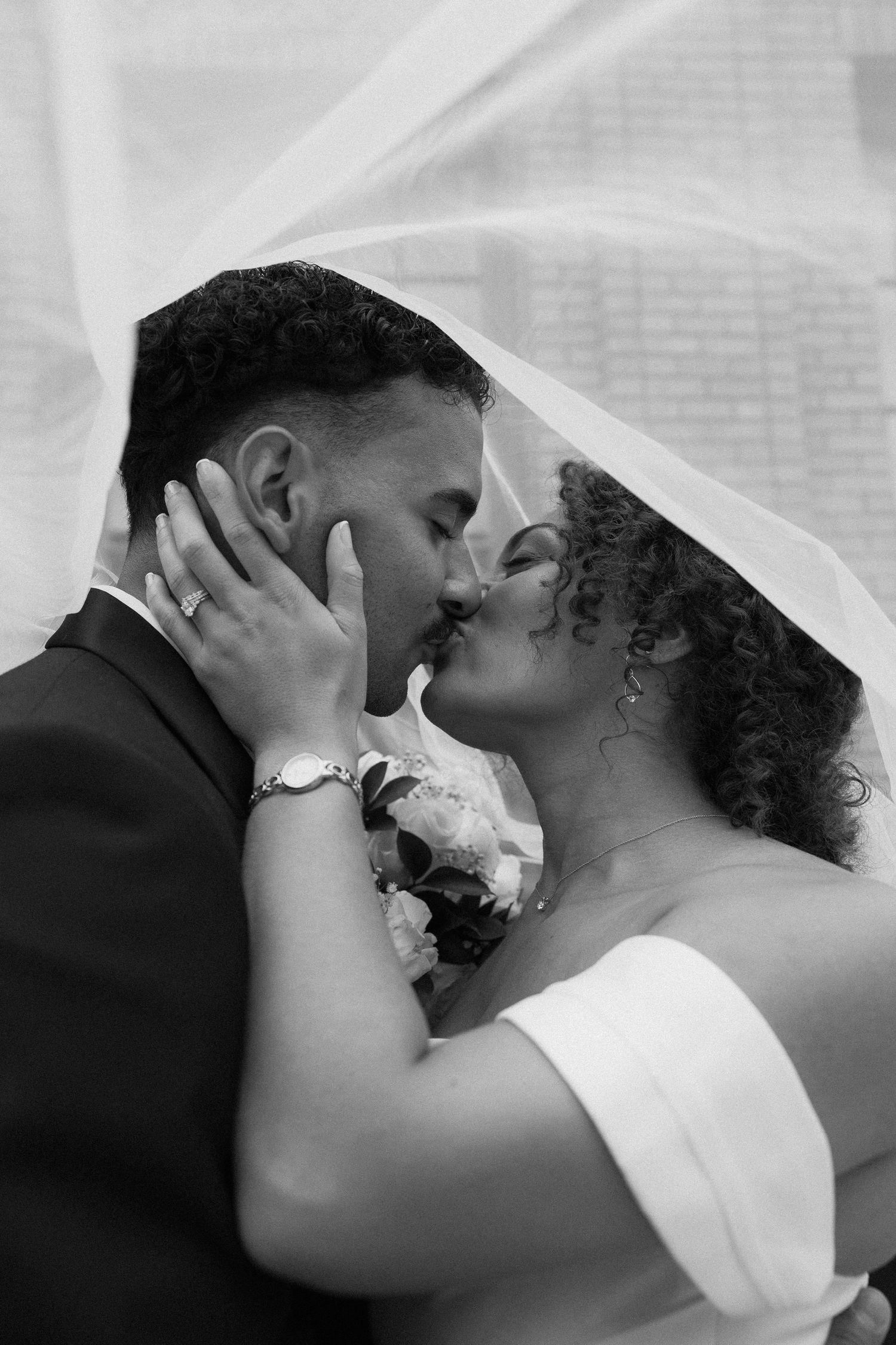 Black and white wedding photo of bride and groom kissing under veil, bride holding bouquet.