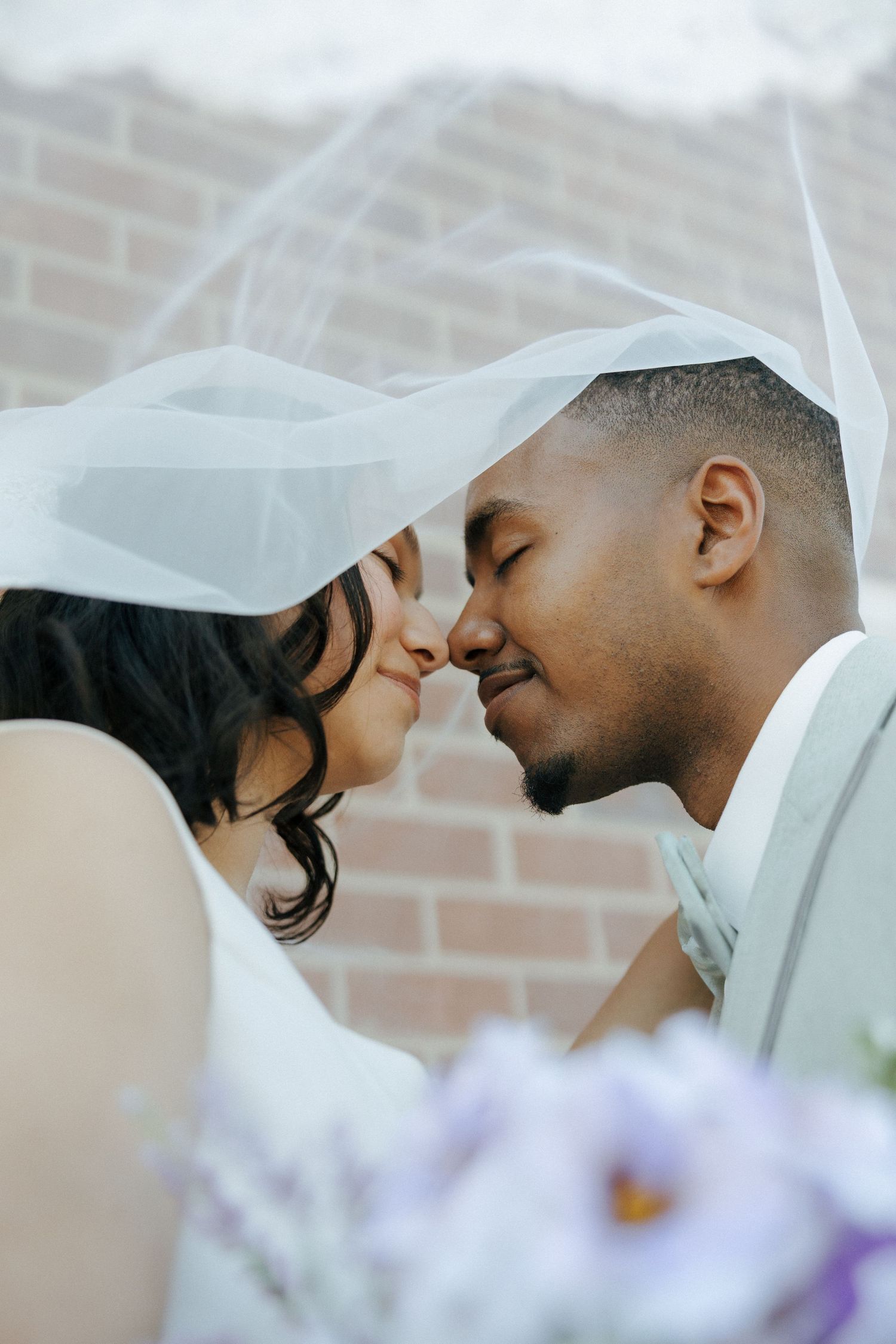 Interracial newlywed couple sharing a kiss under a white wedding veil against a brick wall.