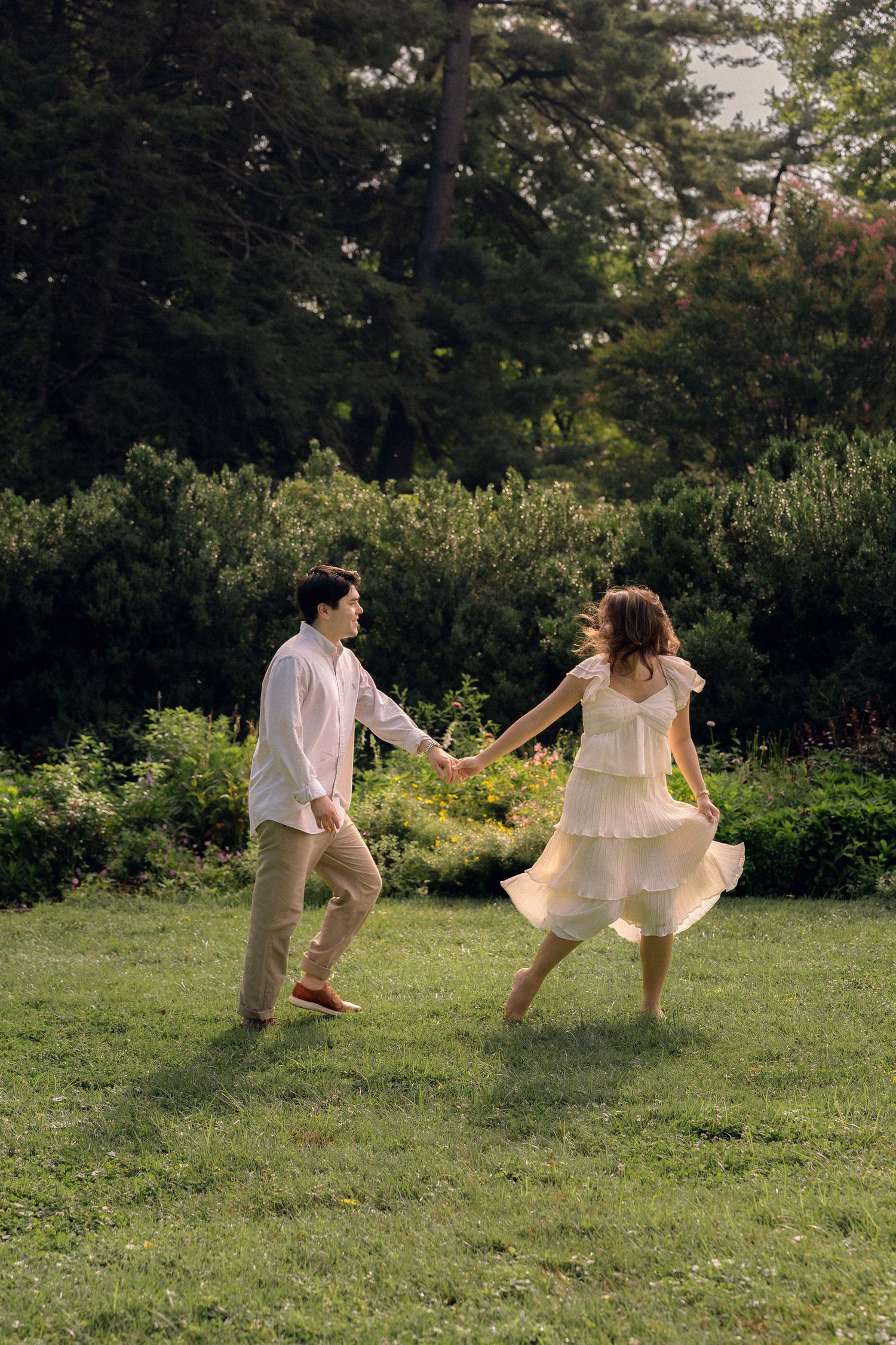 A couple holds hands and dances joyfully on a green lawn surrounded by lush trees and shrubs.
