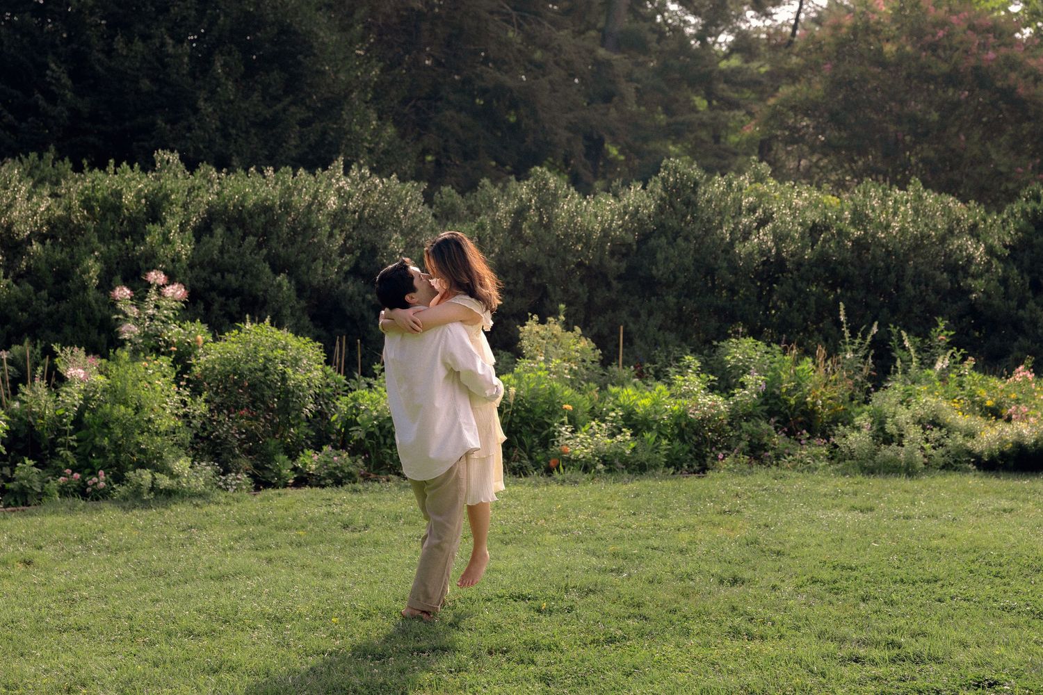 Couple embracing and spinning in a lush green garden surrounded by blooming flowers and trees.
