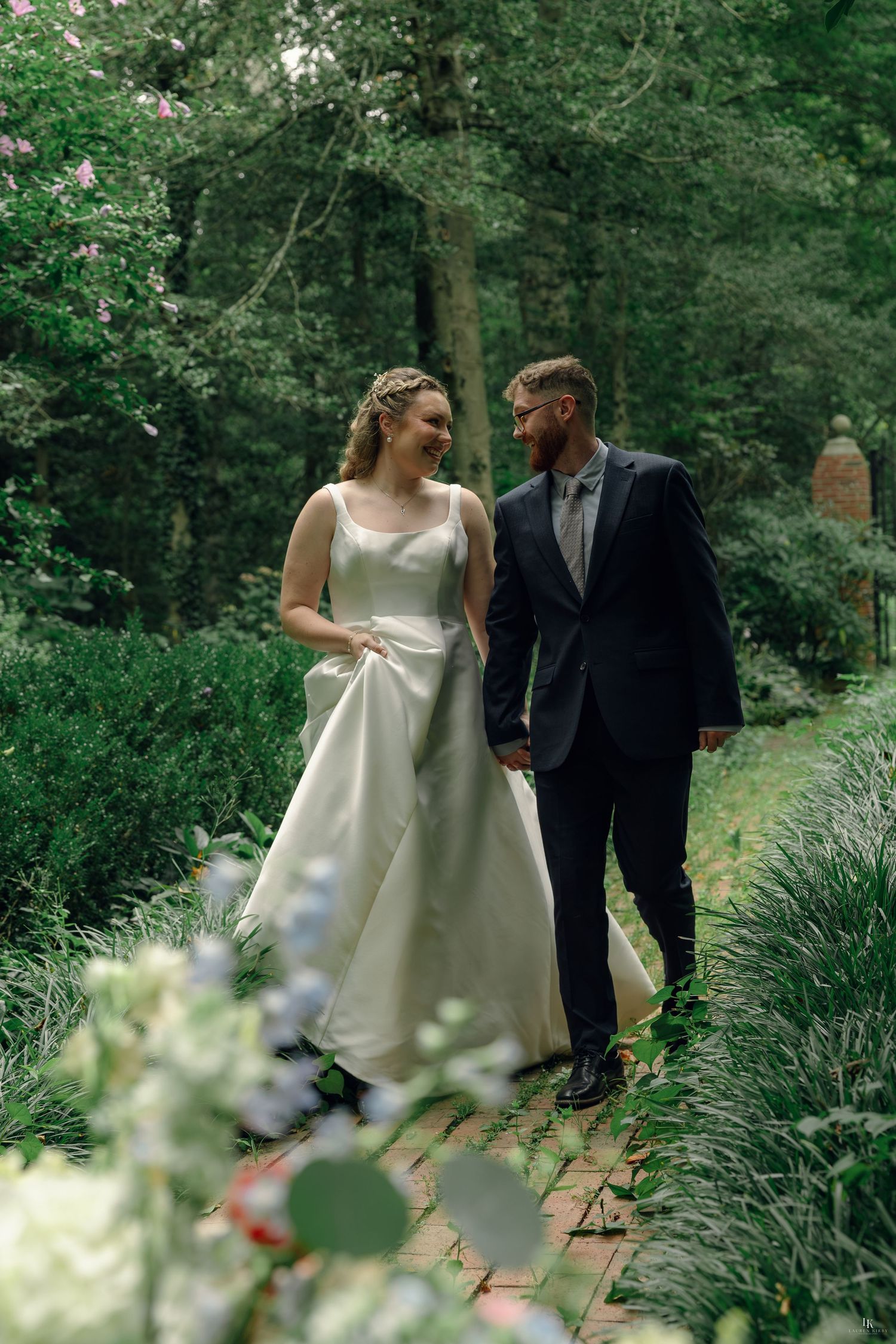 A bride and groom walk hand in hand along a brick path surrounded by lush greenery on their wedding day.