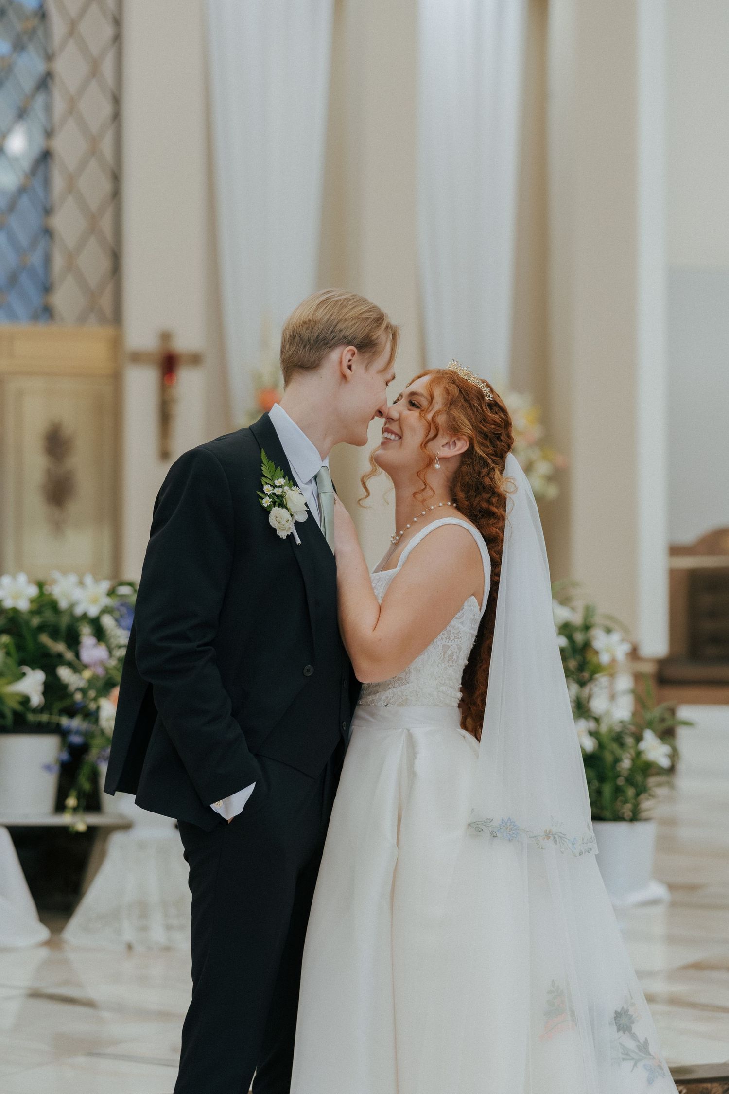 Bride and groom sharing a joyful almost-kiss inside a sunlit church adorned with white floral arrangements.