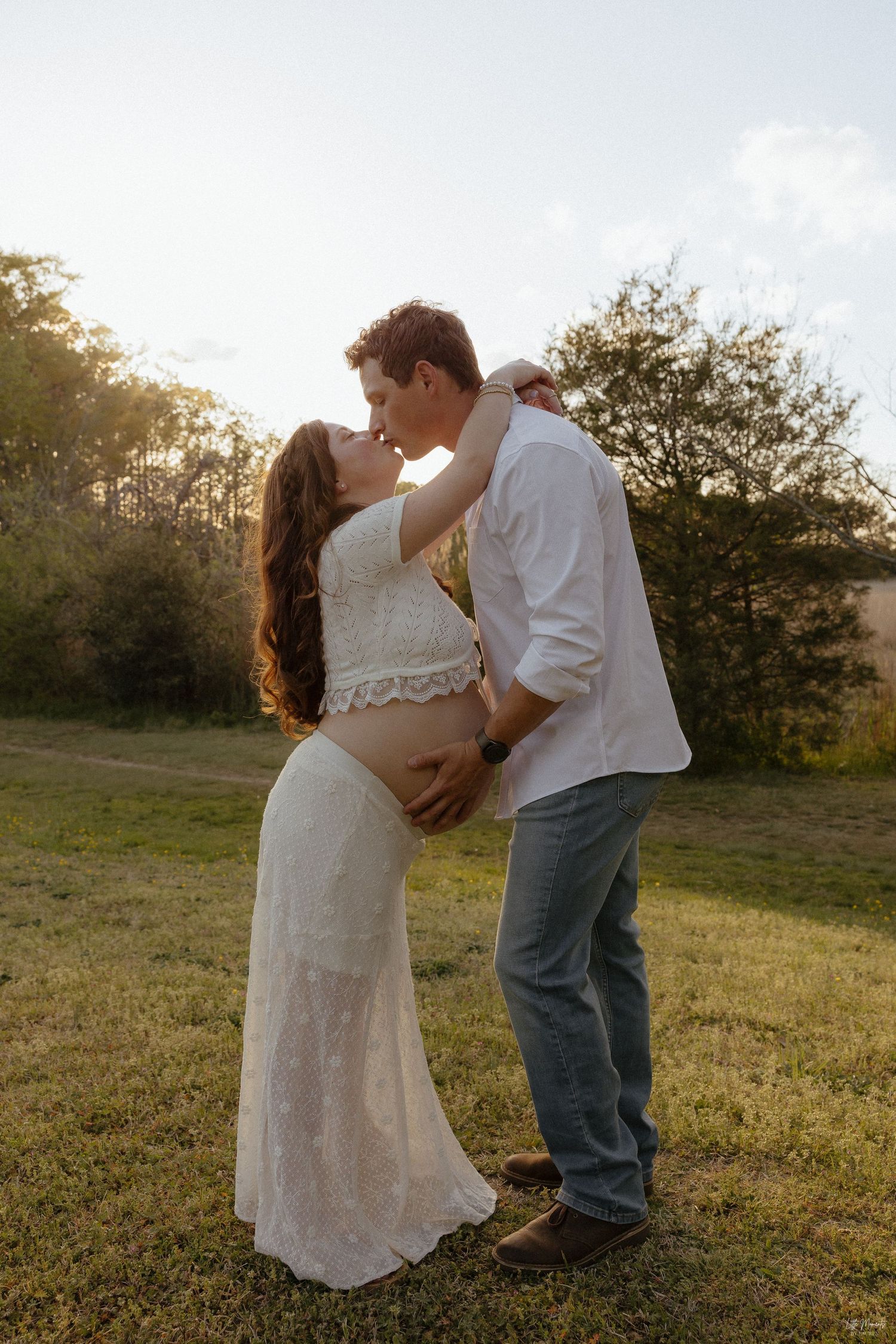 Pregnant couple sharing a tender kiss at golden hour in a lush green outdoor setting.