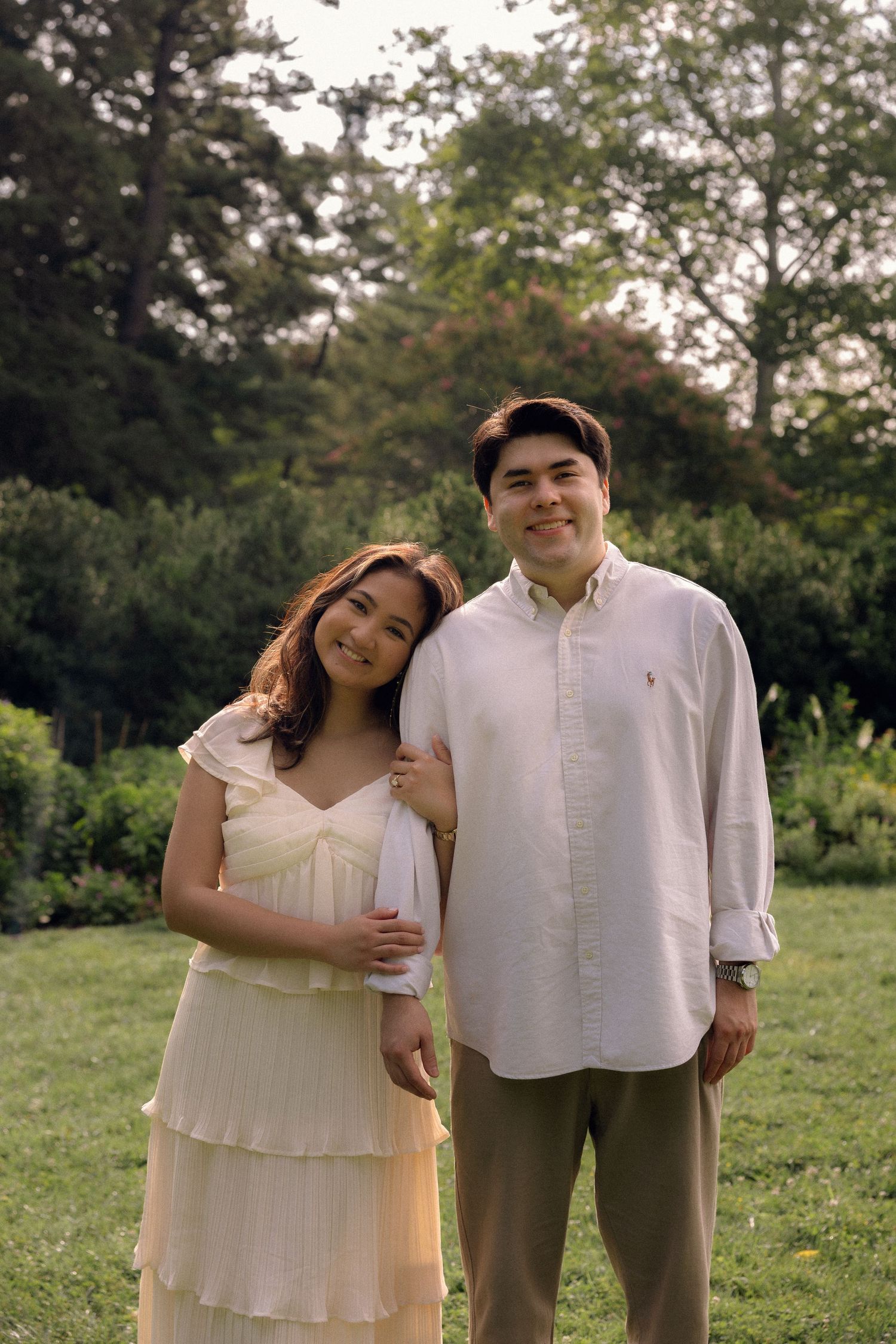 A smiling couple poses together in a lush green garden, woman in white dress, man in light button-up shirt.