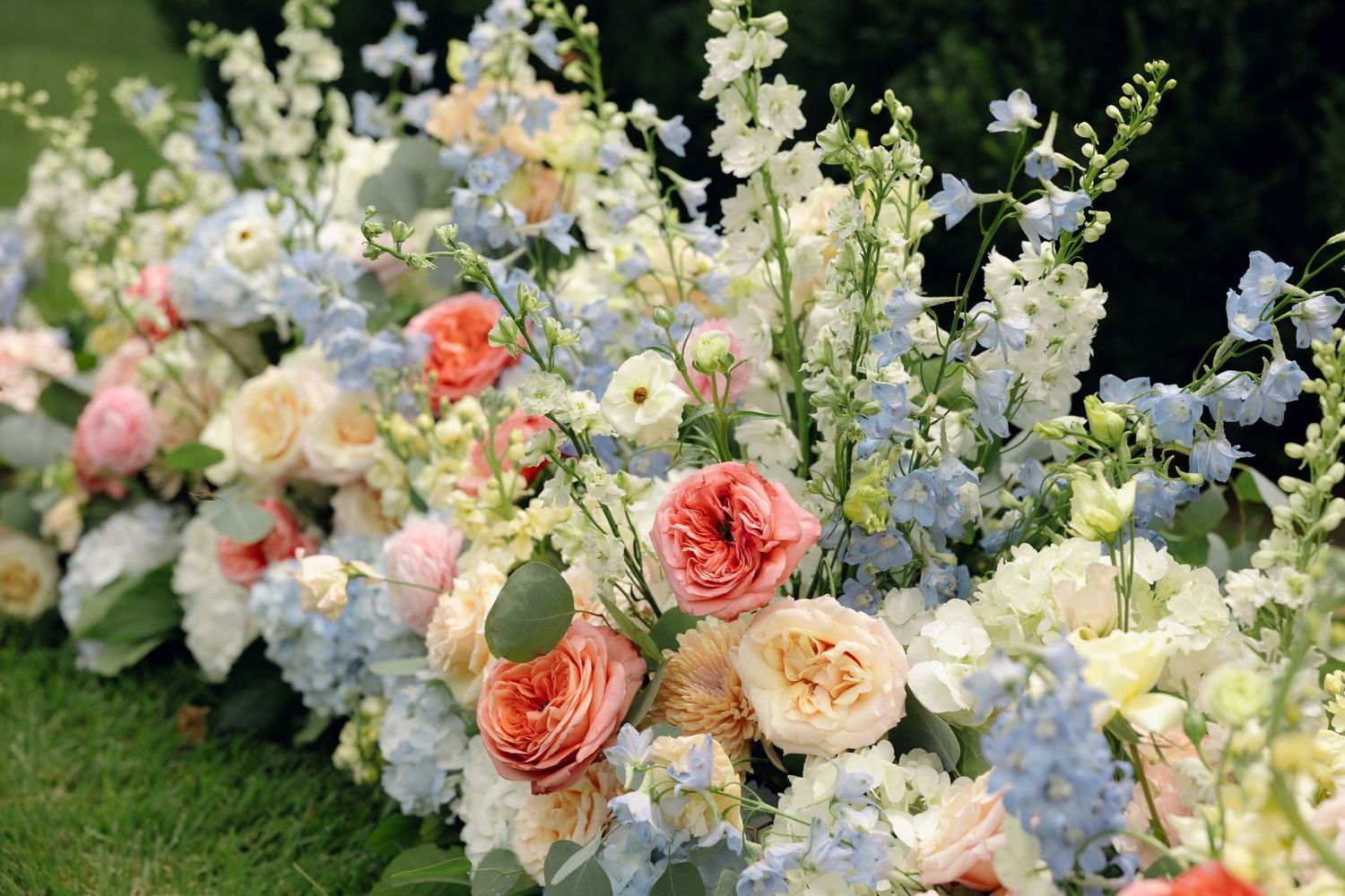 Lush wedding floral arrangement featuring coral garden roses, blue delphiniums, and cream blooms on green grass.