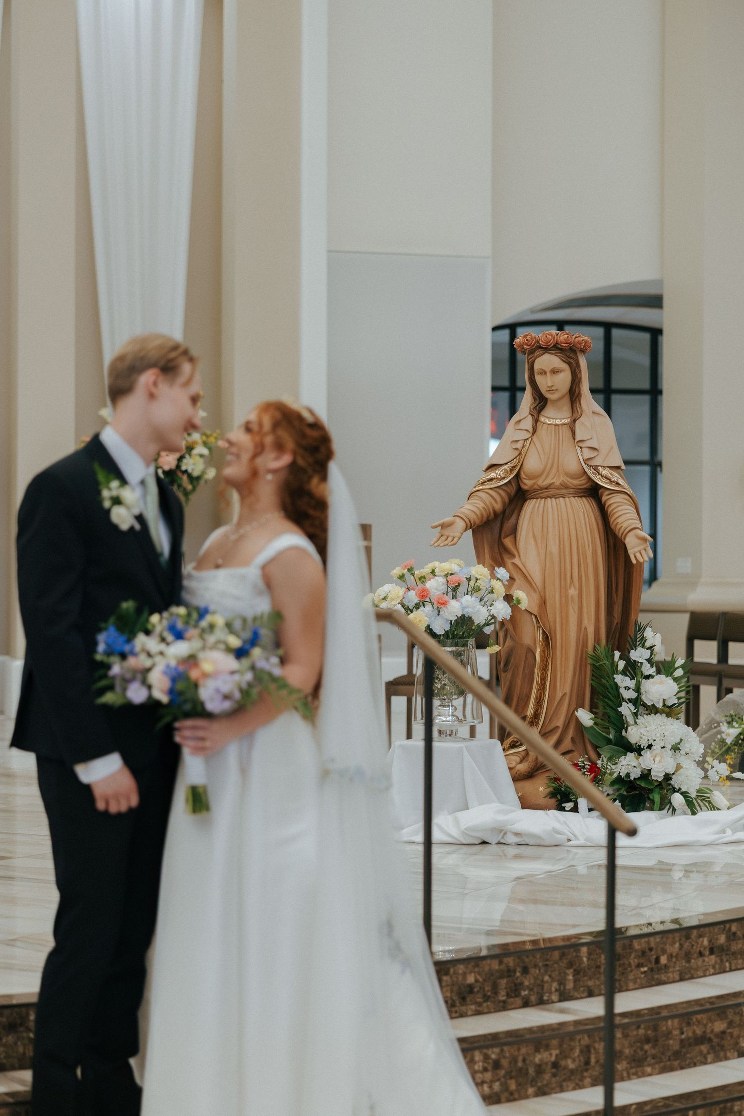 A bride and groom share a romantic moment in a church with a Virgin Mary statue and floral arrangements in the background.