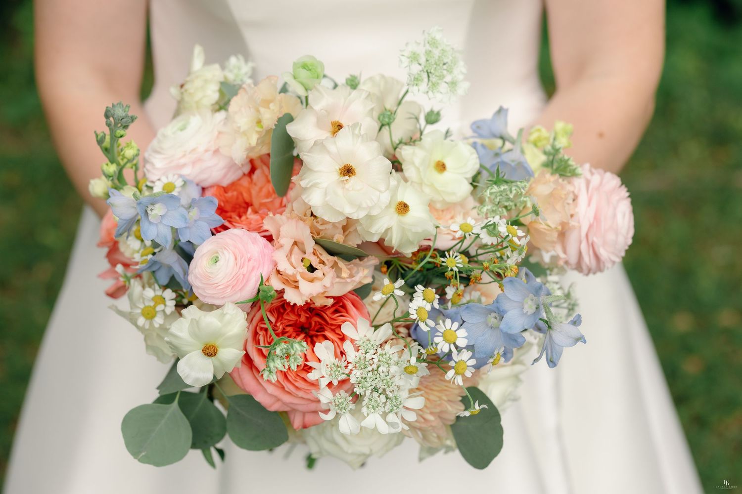 Bride holding a lush wedding bouquet with coral, pink, white, and blue flowers including roses, daisies, and hydrangeas.