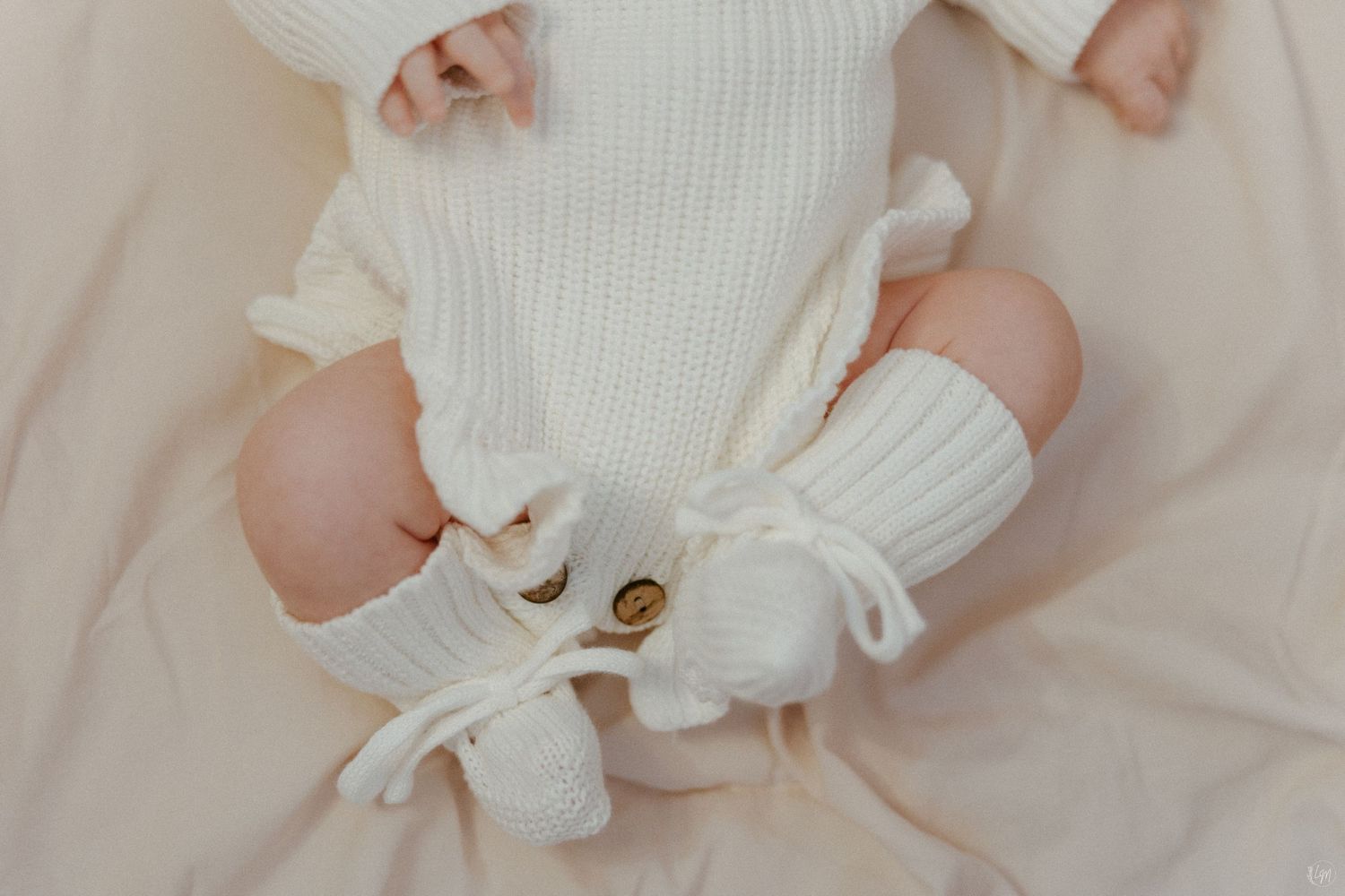 Newborn baby in white knit romper and booties lying on soft cream blanket, cozy infant outfit.