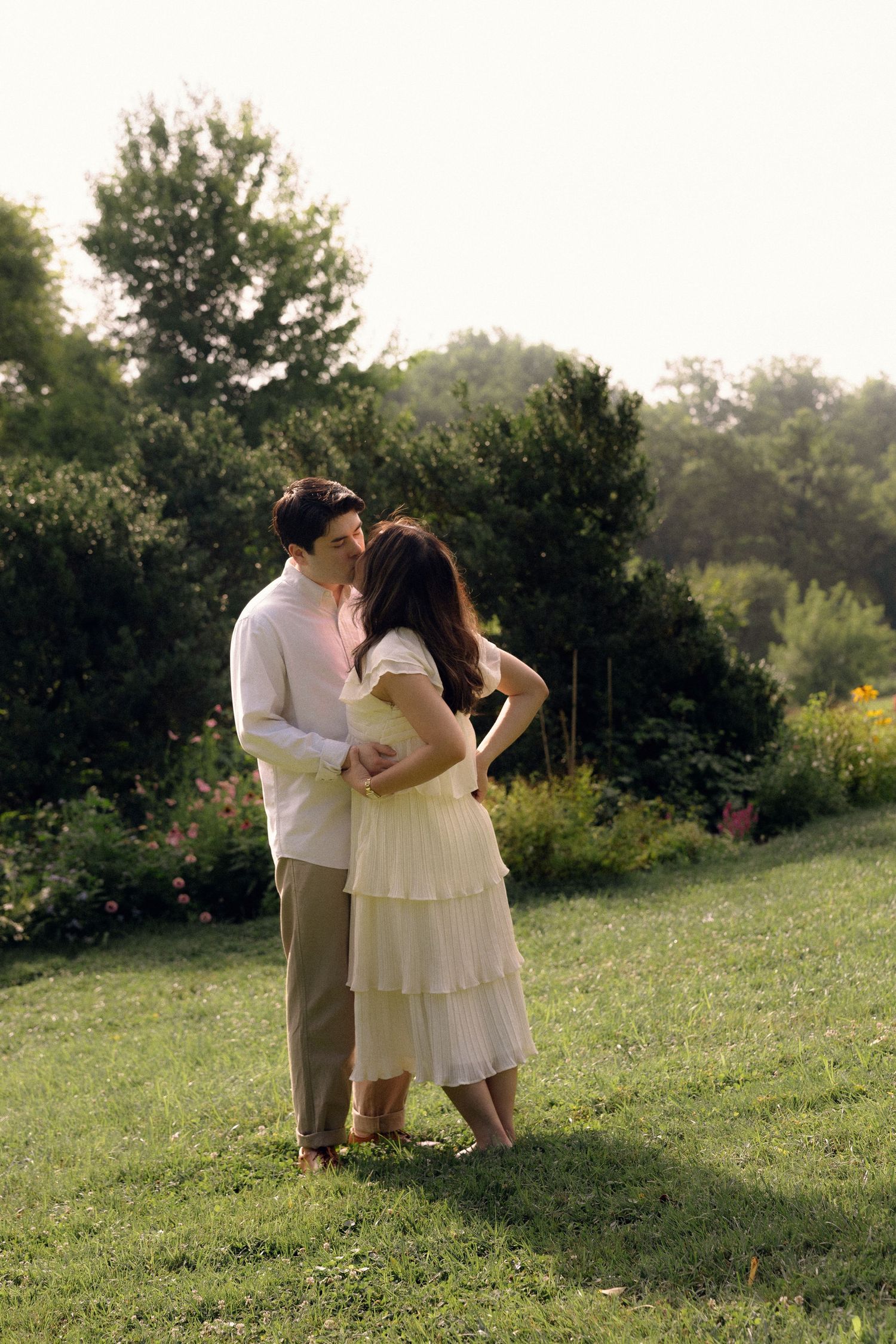 A couple embraces and kisses in a lush green garden at golden hour, woman in white dress, man in white shirt.