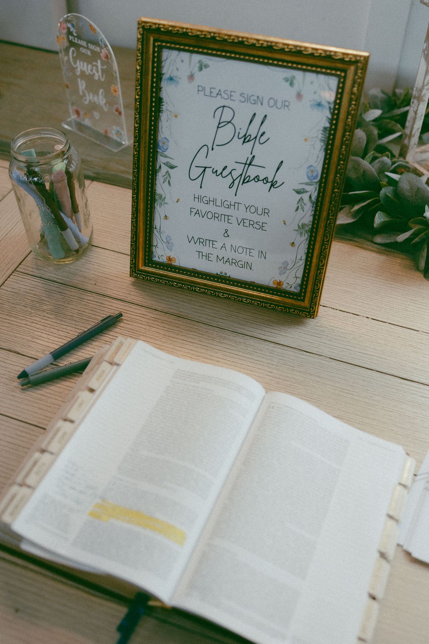 Bible guestbook display at wedding with open highlighted Bible, pens, and framed sign asking guests to write favorite verses.