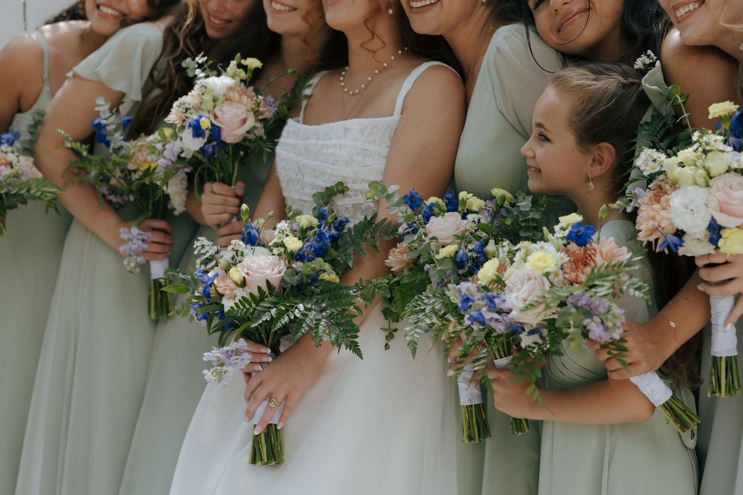 Bride and bridesmaids in sage green dresses holding colorful bouquets with blue, white, and peach flowers at a wedding.