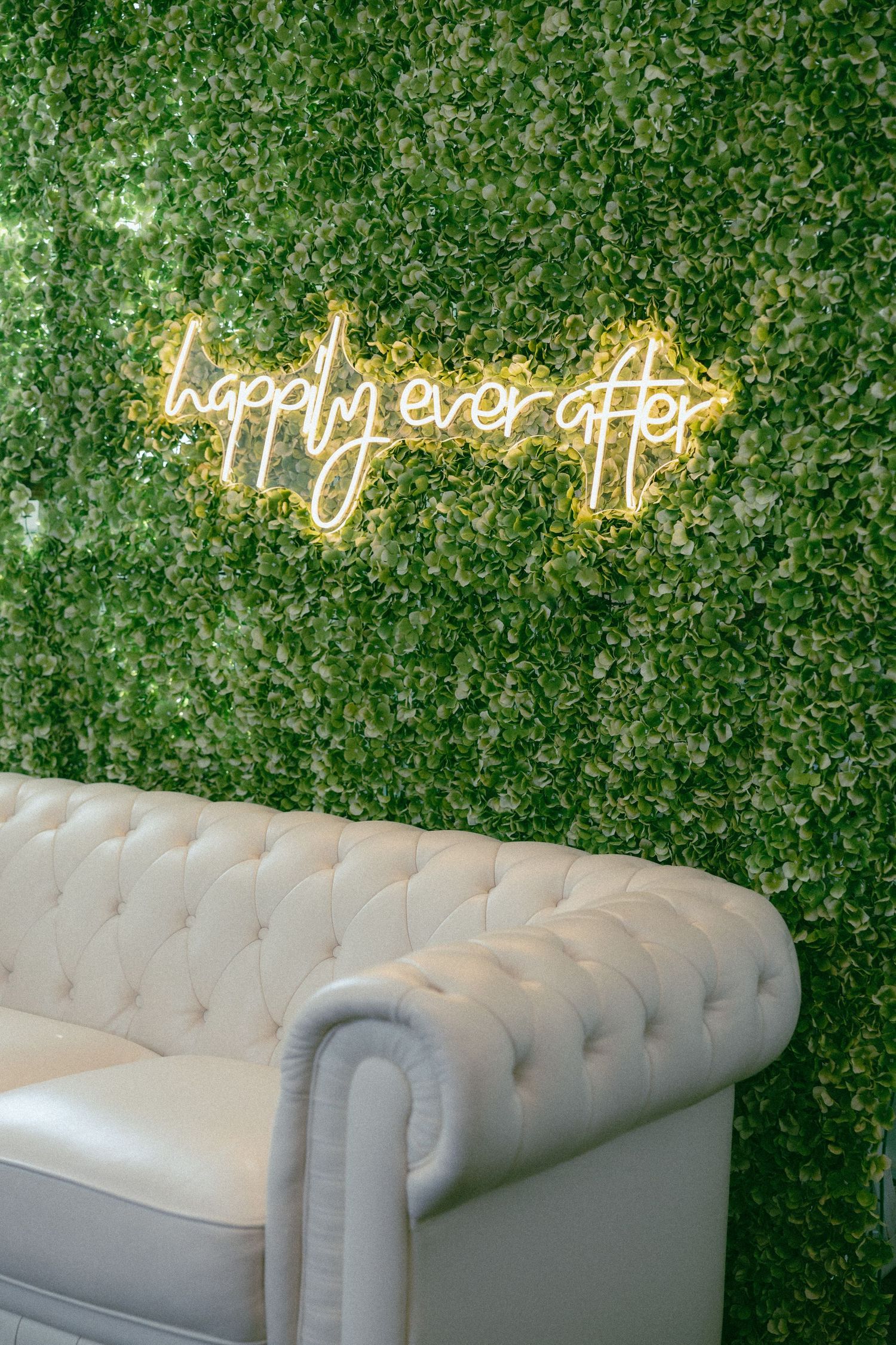 Neon happily ever after sign glowing on a green hedge wall above a white tufted sofa.
