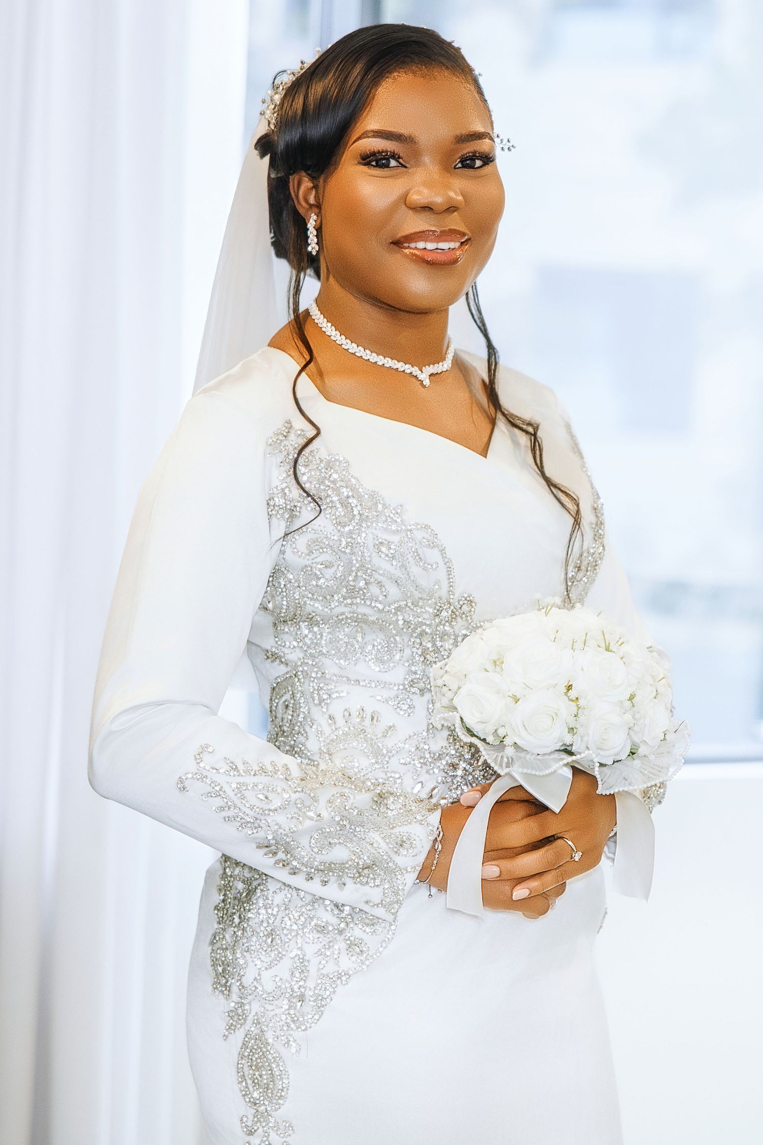 Elegant Black bride in white long-sleeve lace wedding gown holding white bouquet, wearing veil and diamond jewelry.