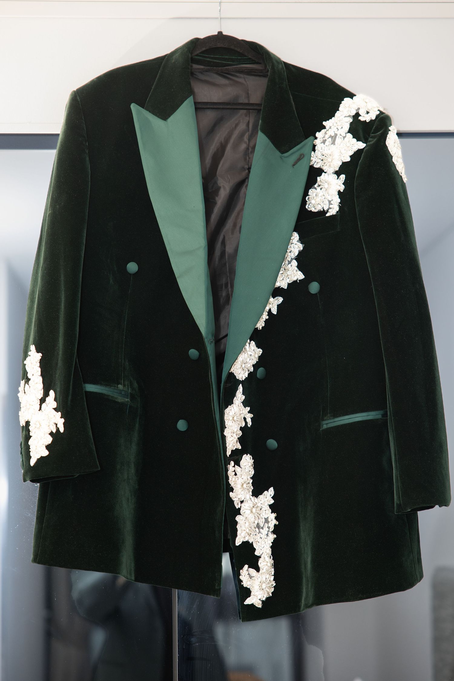 A luxurious dark green velvet double-breasted blazer with white floral lace applique details on a hanger.