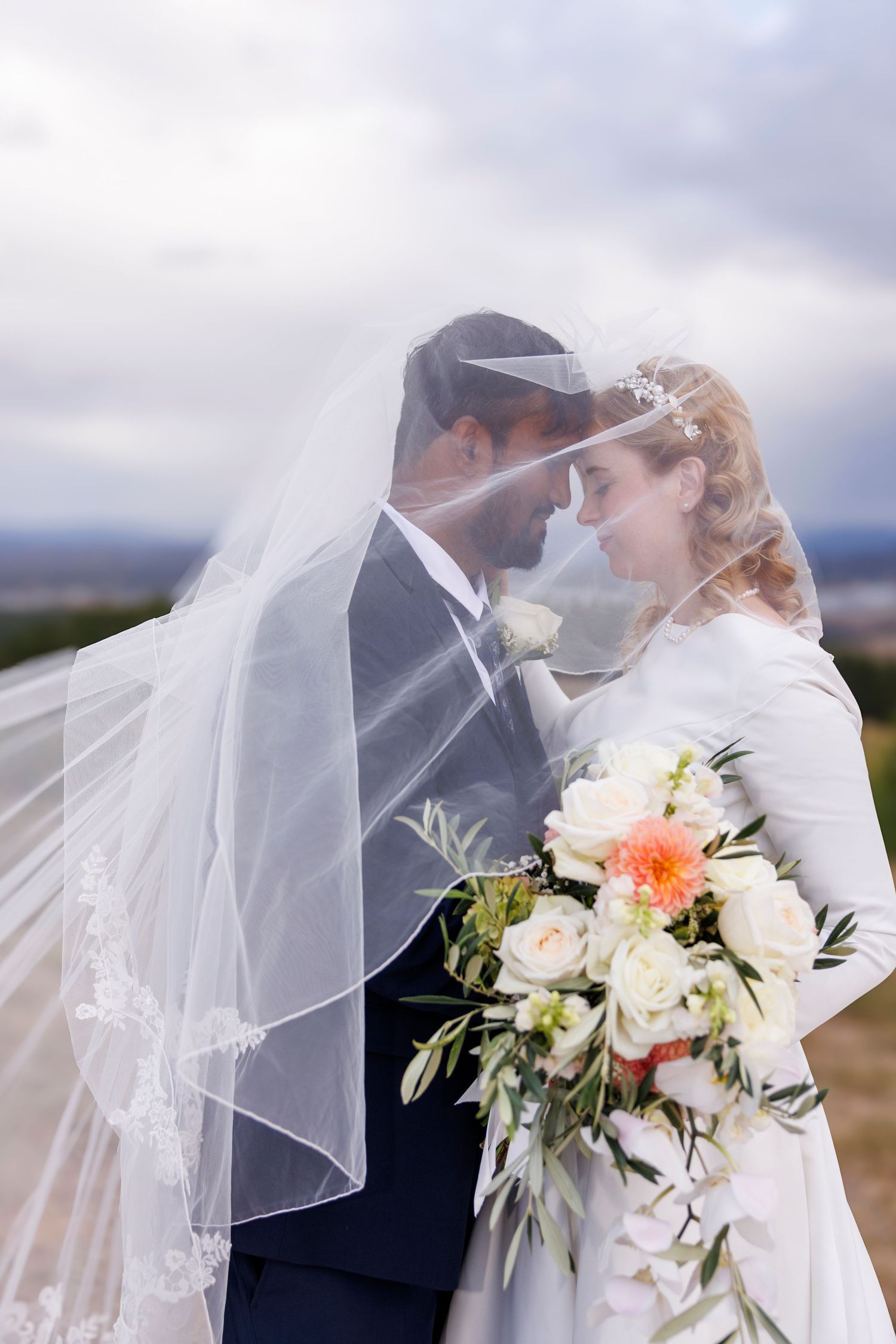 Bride and groom sharing an intimate moment under flowing wedding veil outdoors.