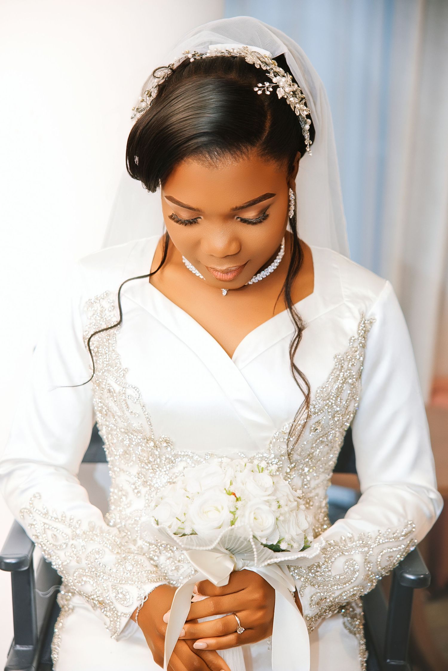 Elegant Black bride in white beaded gown with veil and floral headpiece holds white rose bouquet while looking down.