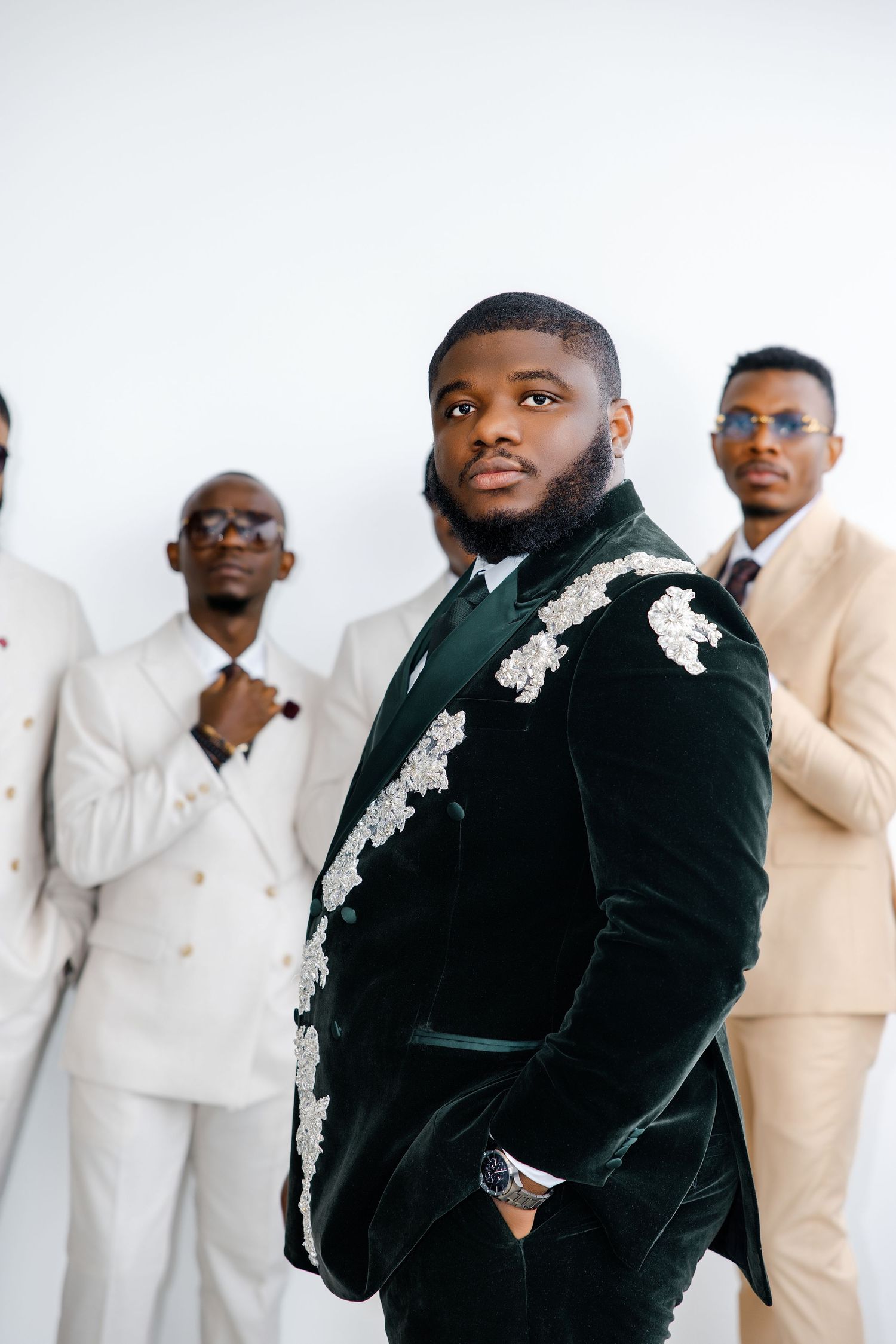 A groom in a green velvet embroidered suit stands confidently with groomsmen in beige suits behind him.