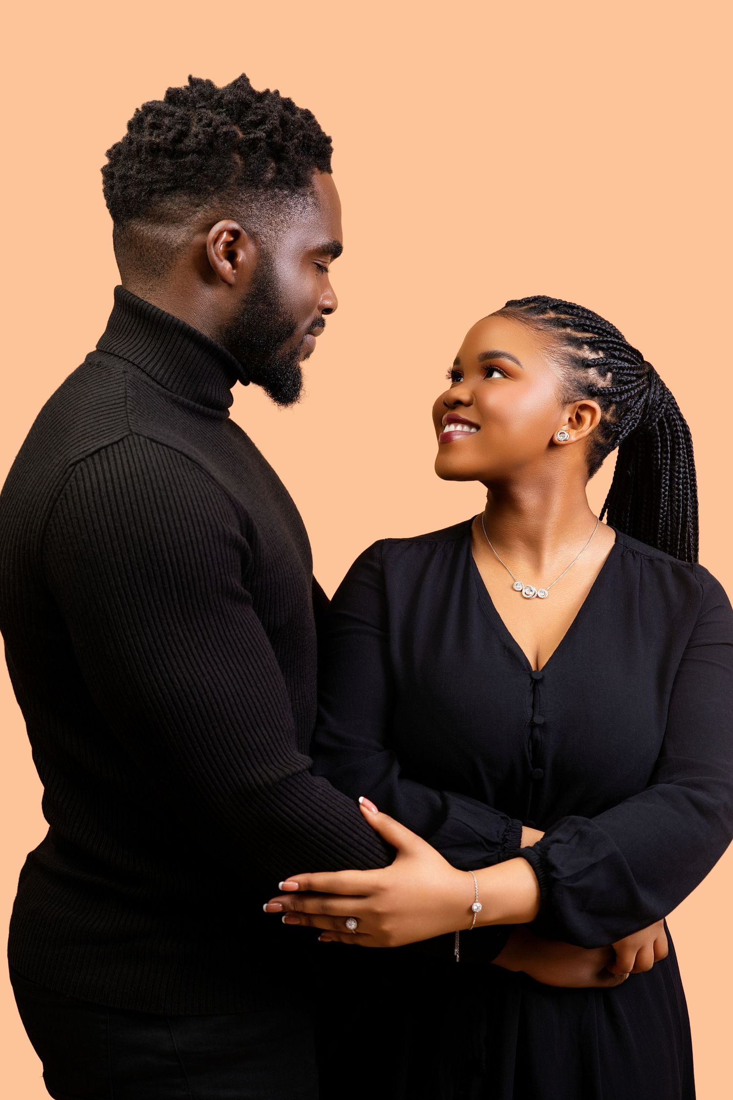 Black couple in black outfits sharing a tender moment, looking at each other against a beige backdrop.