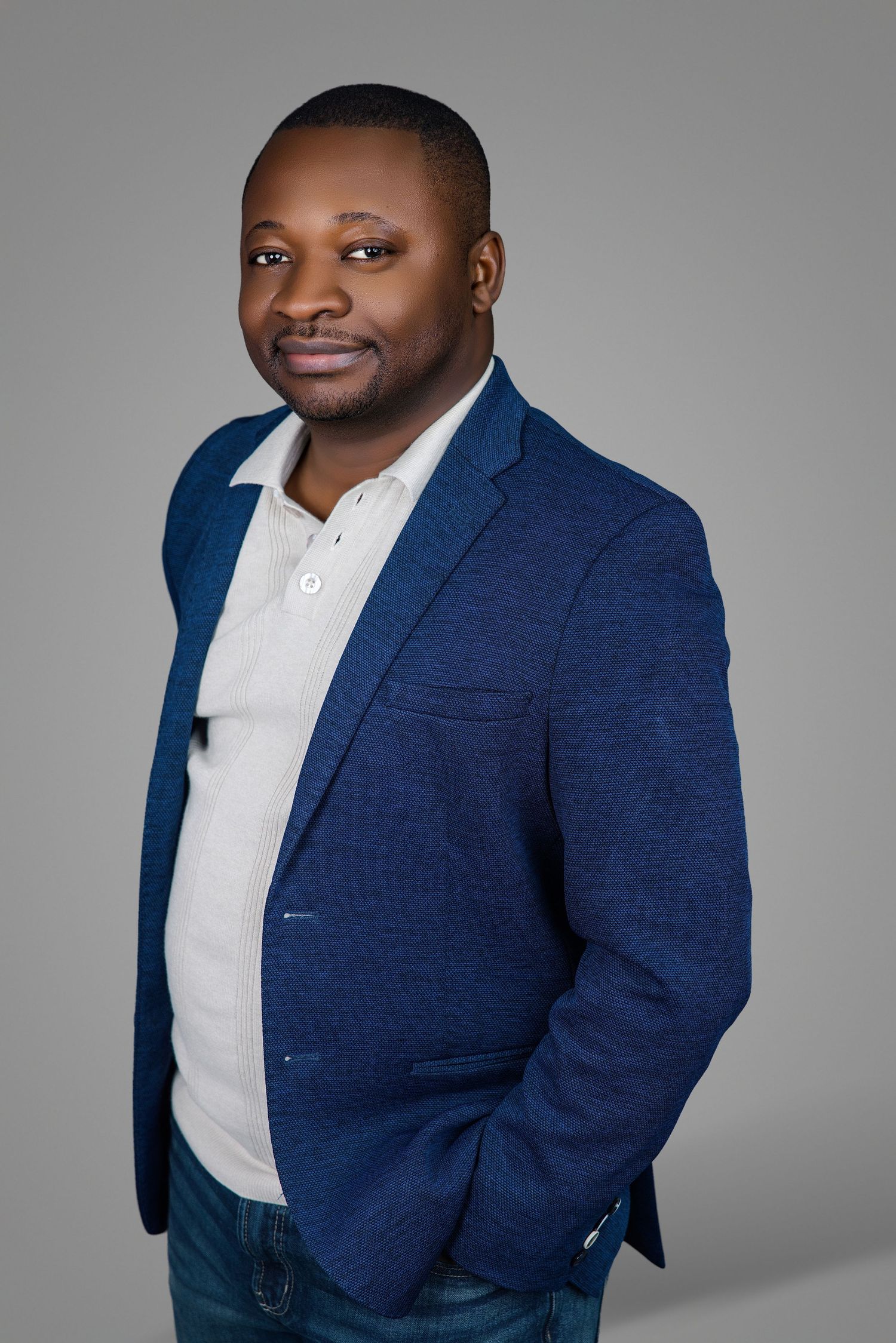 A man in a navy blazer and white polo poses with hands behind his back in a studio photo.