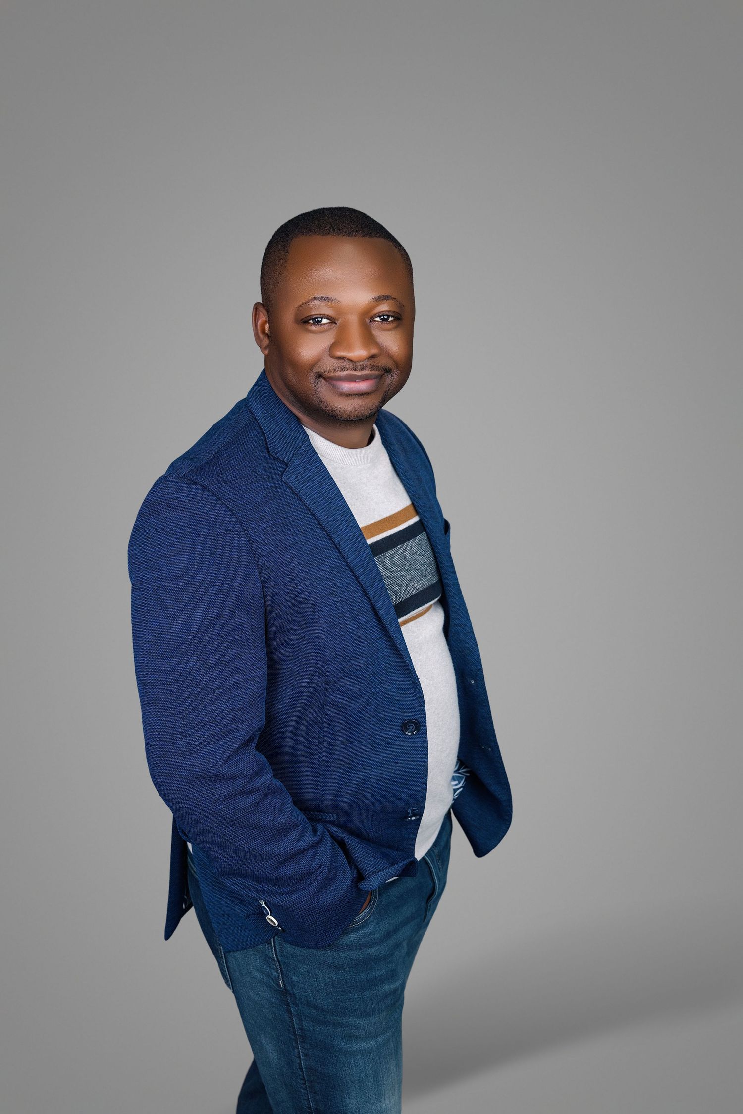 Smiling Black man in blue blazer and striped shirt poses with hands in pockets on gray background.