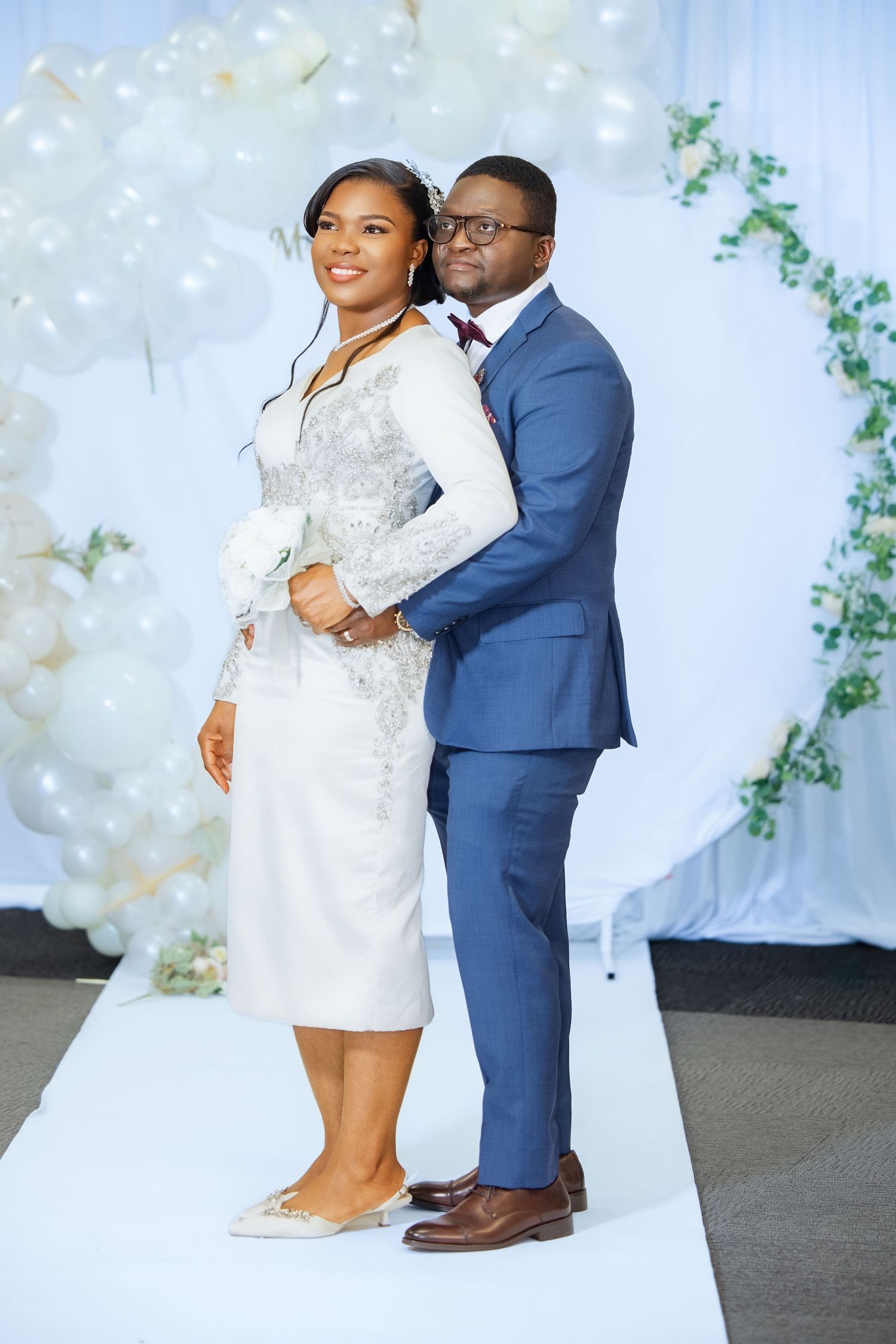 A couple poses elegantly at a wedding, the bride in white lace and groom in a blue suit before a balloon arch.