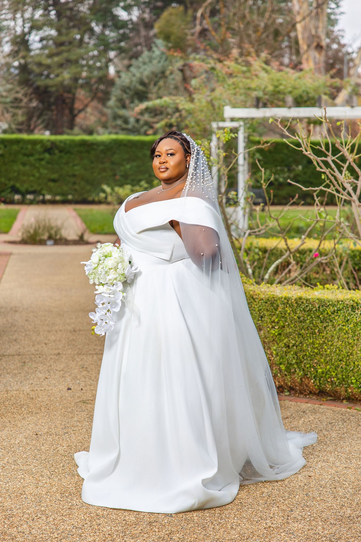 Plus-size bride in elegant off-shoulder white ball gown with pearl veil holding white floral bouquet in garden.