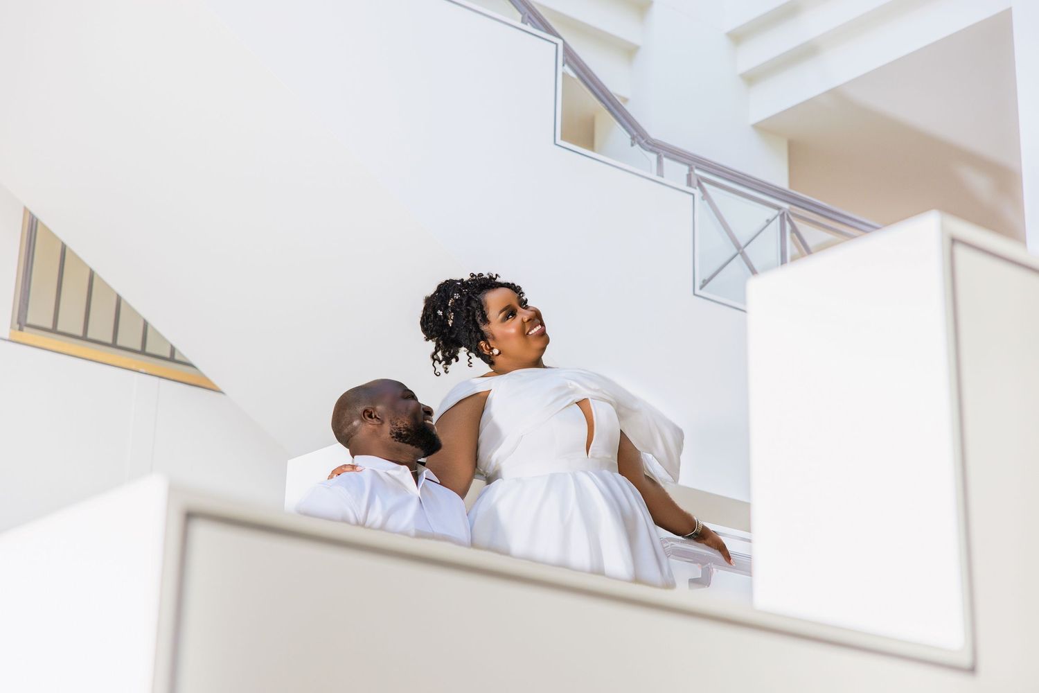 Couple in white wedding attire sharing intimate moment on modern white staircase viewed from below.