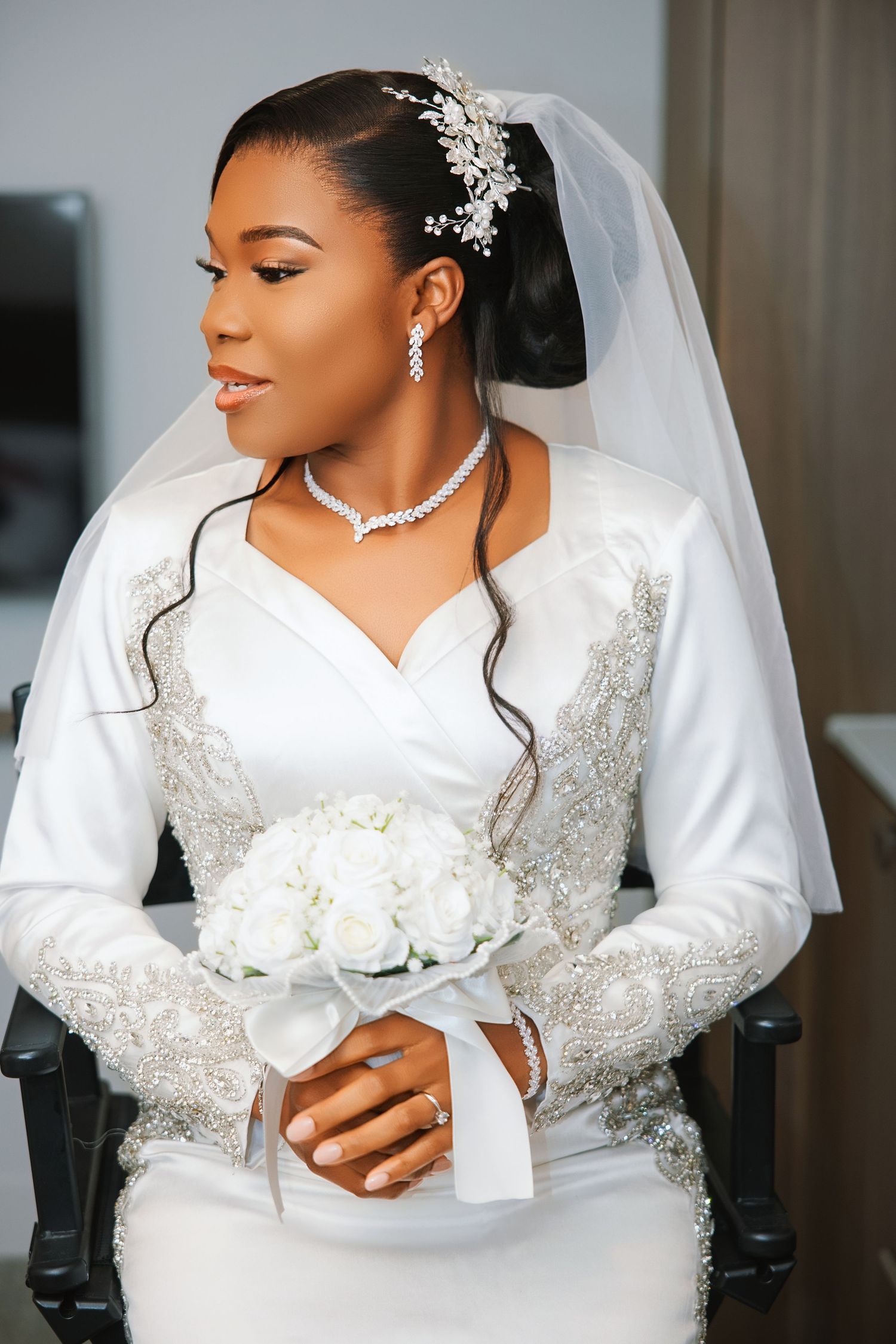 Elegant Black bride in white beaded long-sleeve gown, veil, and diamond jewelry, holding white rose bouquet while seated.