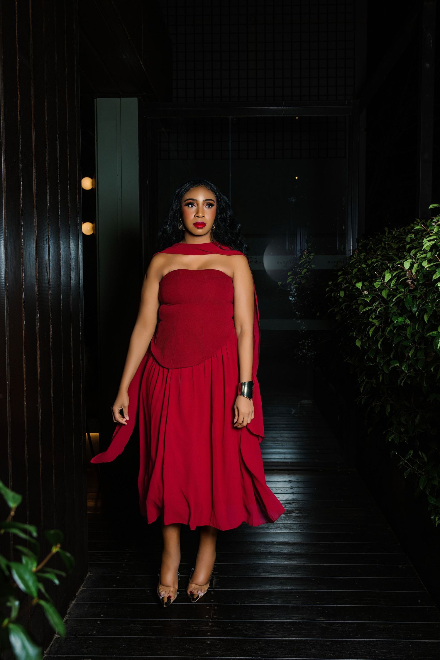 Woman in elegant red strapless midi dress with flowing skirt poses in dark corridor with green plants.