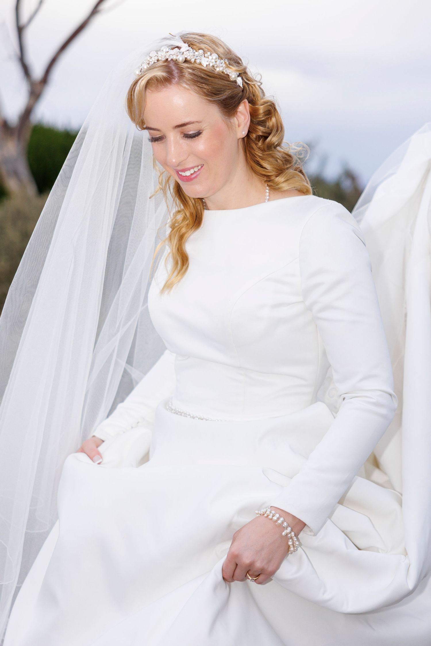 Elegant bride in white long-sleeve gown with tiara and veil smiling outdoors.