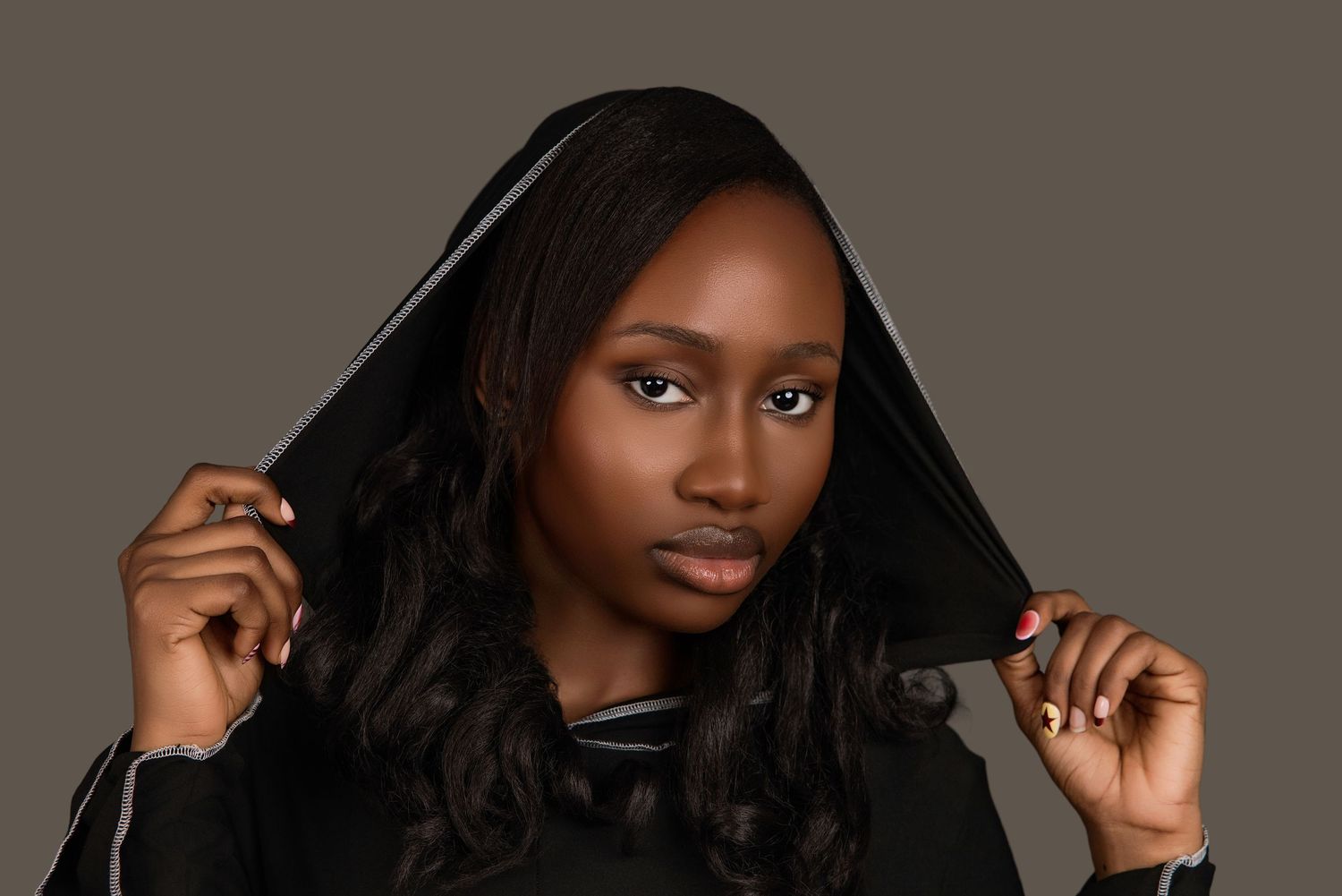 Young Black woman in black hooded outfit posing confidently against a gray background.