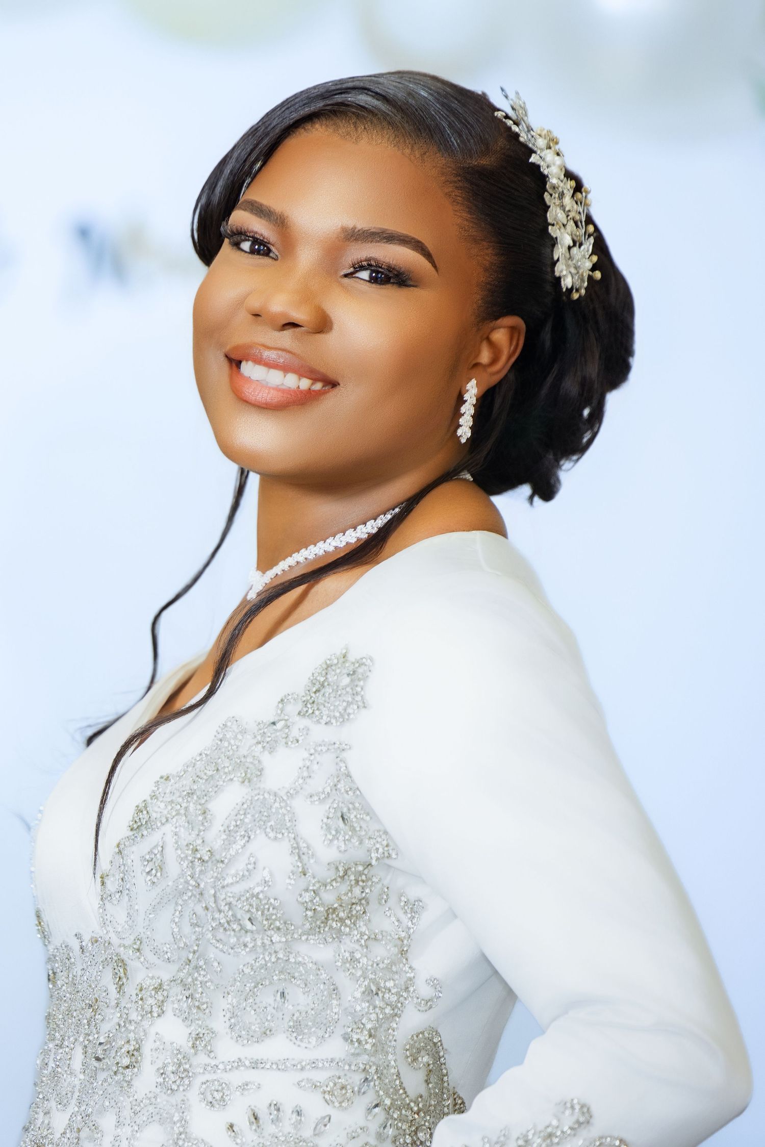 Smiling bride in white embroidered gown with pearl necklace and floral hair accessory at a wedding.