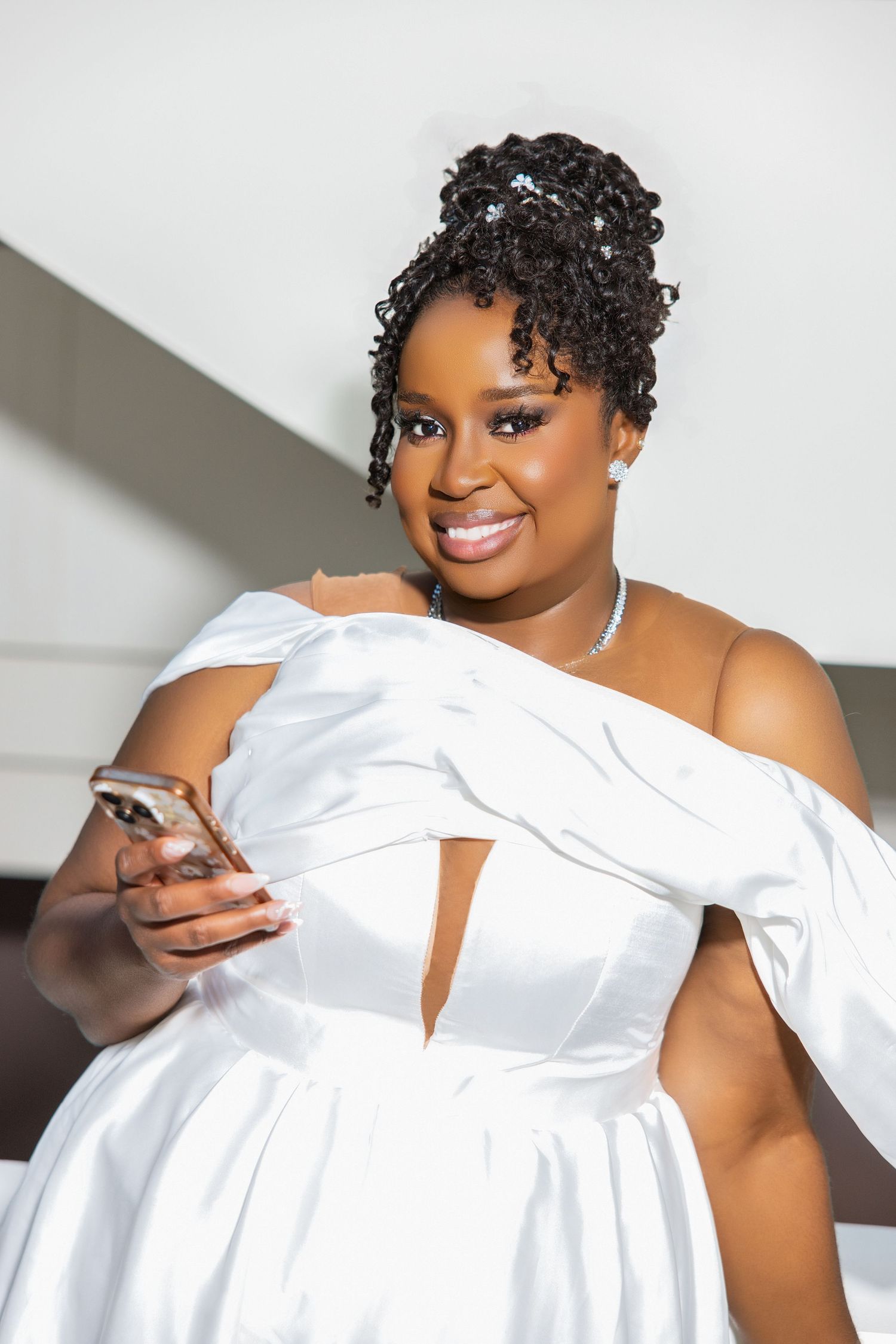 Radiant bride with curly updo in white off-shoulder satin dress holding smartphone on modern staircase.