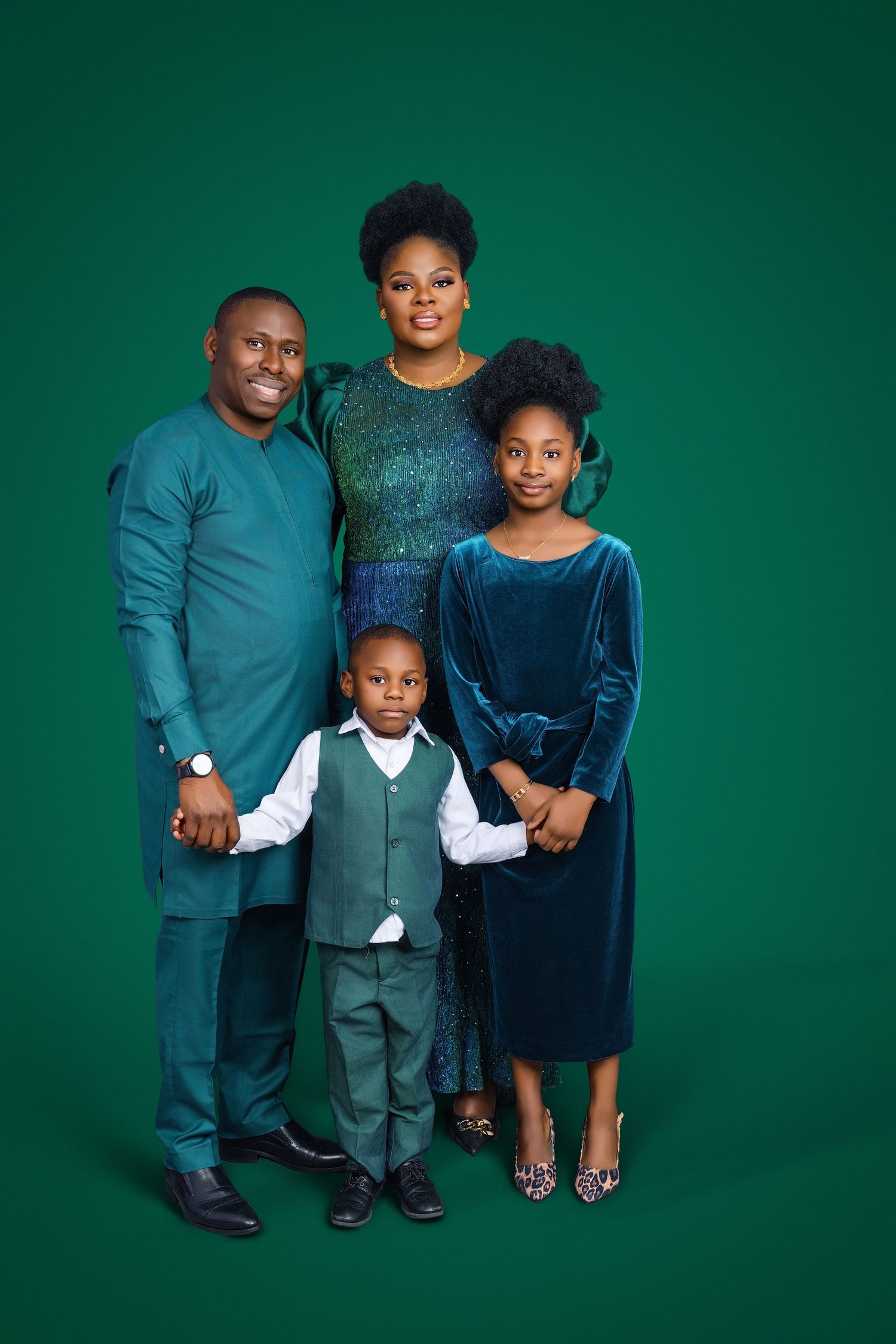 Happy Black family of four in coordinating teal and green outfits posing together on a green background.